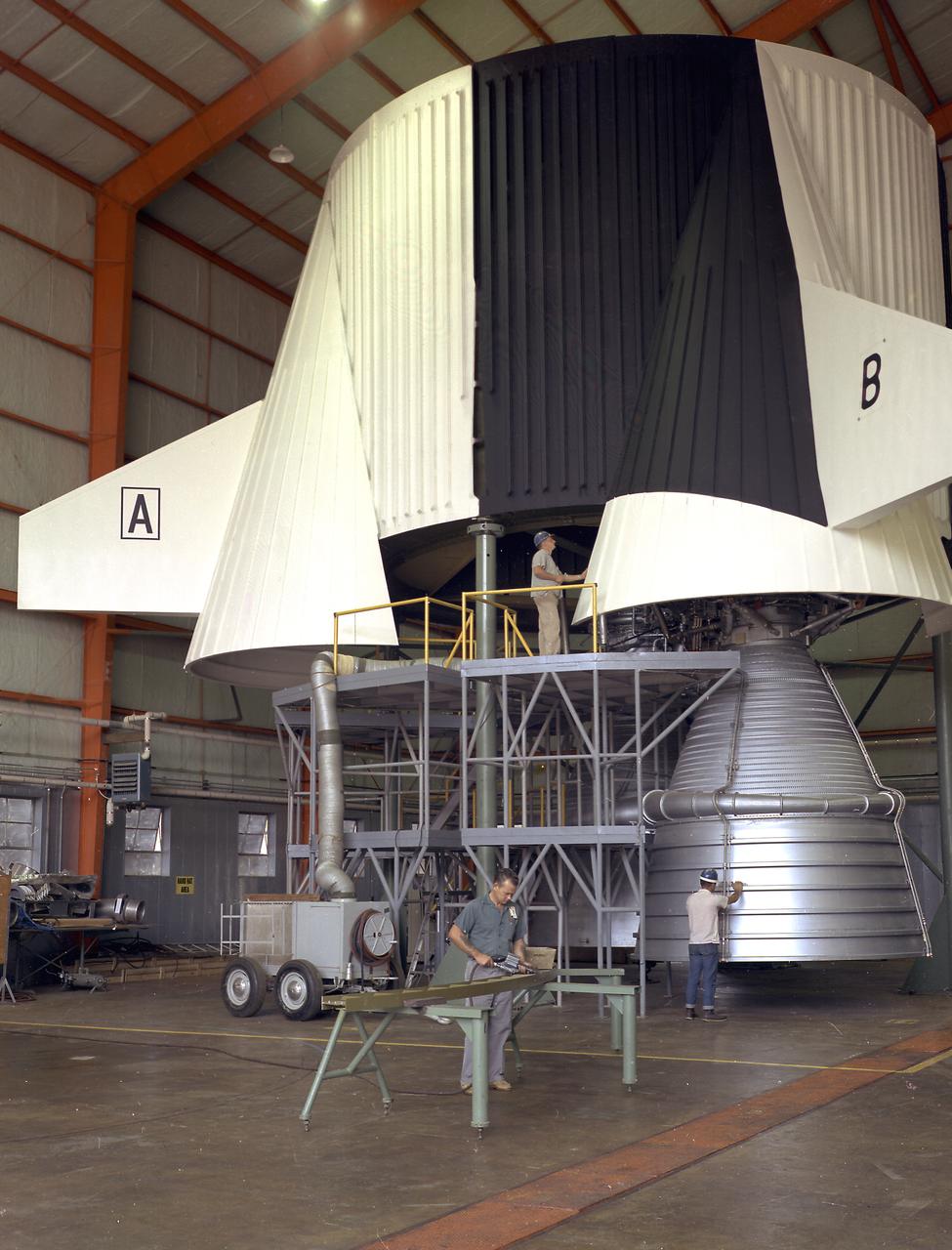 This image illustrates technicians working on a full scale engineering mock-up of a Saturn V S-IC stage thrust structure nearing completion at the Manufacturing Engineering Laboratory at Marshall Space Flight Center. The booster, 33 feet in diameter and 138 feet long, was powered by five F-1 engines that provided 7,500,000 pounds of thrust to start the monstrous vehicle on its journey into space. 
