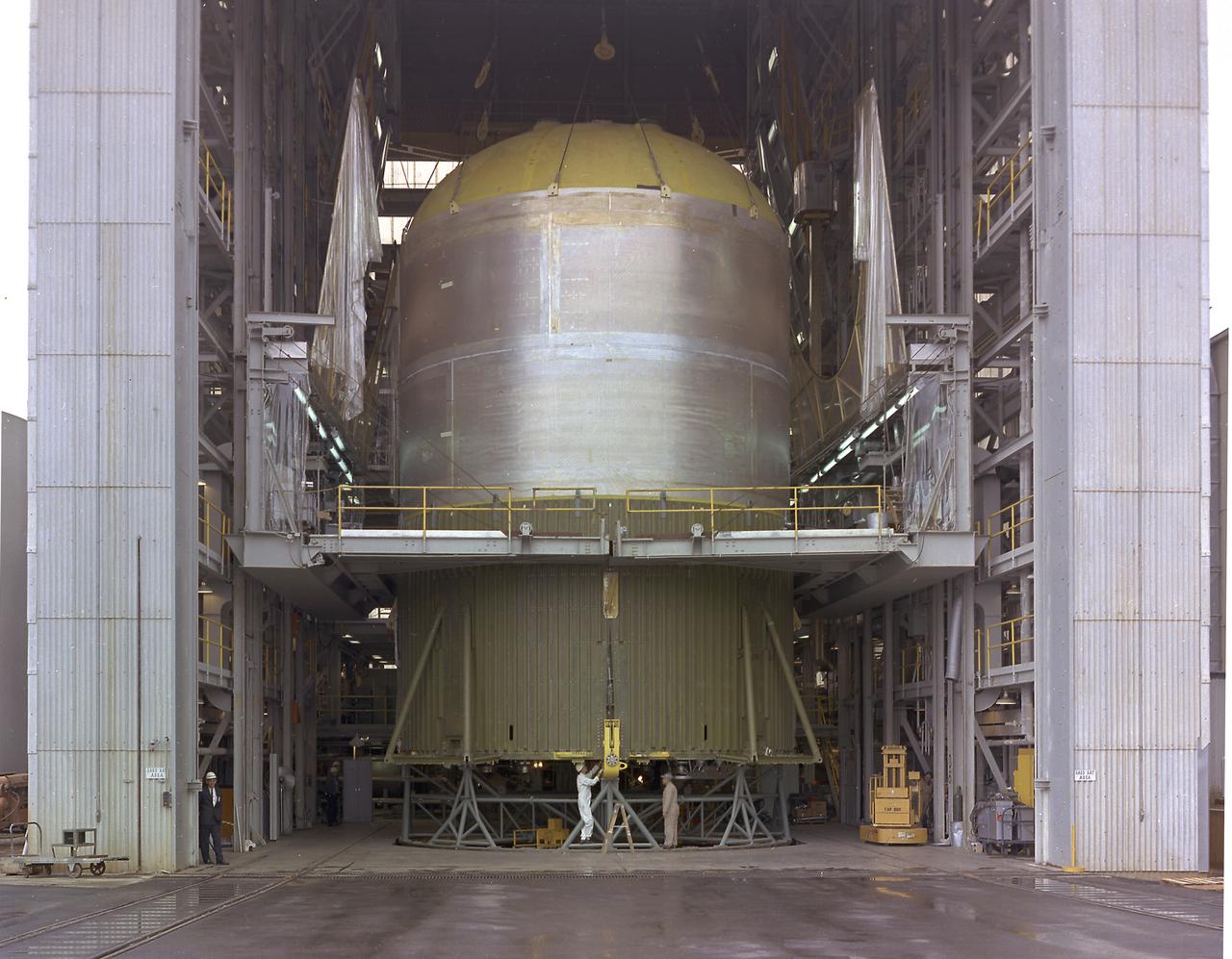This image shows the Saturn V S-IC-T stage (S-IC static test article) fuel tank being attached to the thrust structure in the vehicle assembly building at the Marshall Space Flight Center (MSFC). The S-IC stage utilized five F-1 engines that used liquid oxygen and kerosene as propellant and provided a combined thrust of 7,500,000 pounds. 