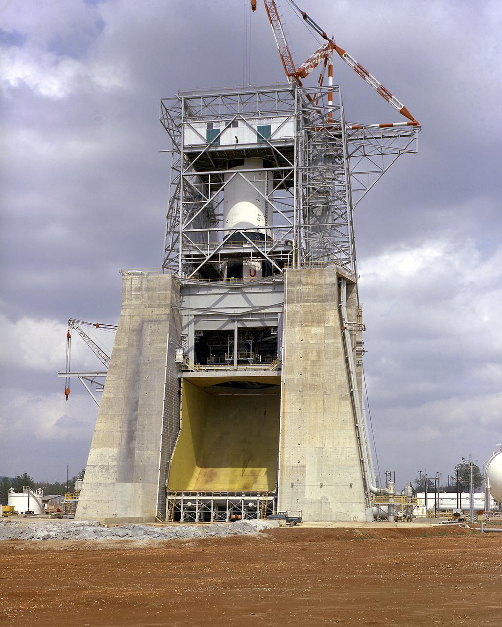 The S-IC-T stage (static firing stage) is installed and awaits the first static firing of all five F-1 engines at the Marshall Space Flight Center S-IC static test stand. Constructed in 1964, the S-IC static test stand was designed and constructed to develop and test the first stage of the Saturn V launch vehicle that used five F-1 engines. Each F-1 engine developed 1,500,000 pounds of thrust for a total liftoff thrust of 7,500,000 pounds. To handle this research and development effort, the stand contains 12,000,000 pounds of concrete on its base legs that are planted down to bedrock 40 feet below ground level. Of concrete and steel construction, the stand foundation walls are 4 feet thick, and topped by a crane with a 135-foot boom. With the boom in the up position, the stand is given an overall height of 405 feet, placing it among the highest structures in Alabama at the time. 