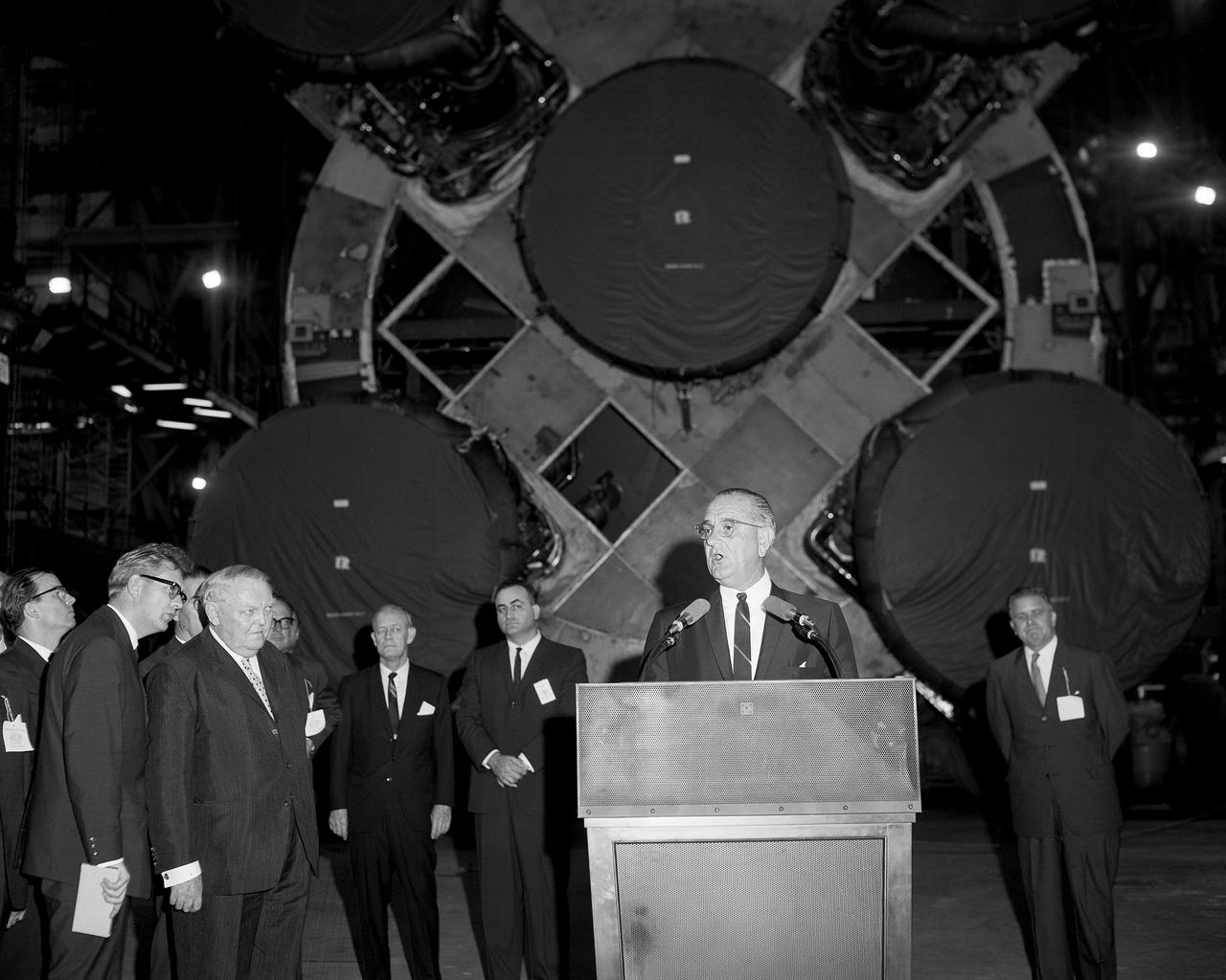 CAPE KENNEDY, Fla. -- At the Kennedy Space Center in Florida, President Lyndon B. Johnson speaks in the Vehicle Assembly Bulling with West German Chancellor Erhard on left, NASAS Administrator James Webb on right, and NASA Director of launch operations, Rocco Petrone, behind President Johnson. In the background is the first stage of a Saturn V rocket which will be used to launch astronauts to the moon as part of the Apollo Program. Photo Credit: NASA
