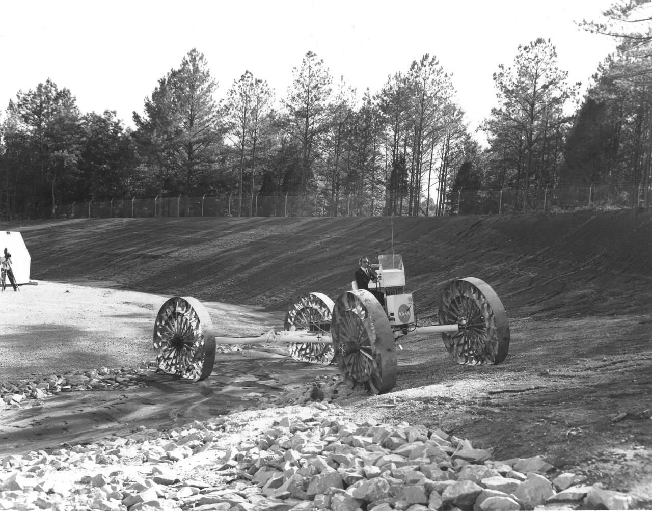 An engineer demonstrates a Mobility Test Article (MTA) at NASA’s Marshall Space Flight Center (MSFC) as he crosses a soft clay strip onto rocky ground. This unit, weighing 1/6th as much as an actual vehicle, was built by the Bendix Corporation and was one of the concepts of a possible Lunar Roving Vehicle (LRV). The data provided by the MTA helped in designing the LRV, developed under the direction of MSFC. The LRV was designed to allow Apollo astronauts a greater range of mobility during lunar exploration missions. 