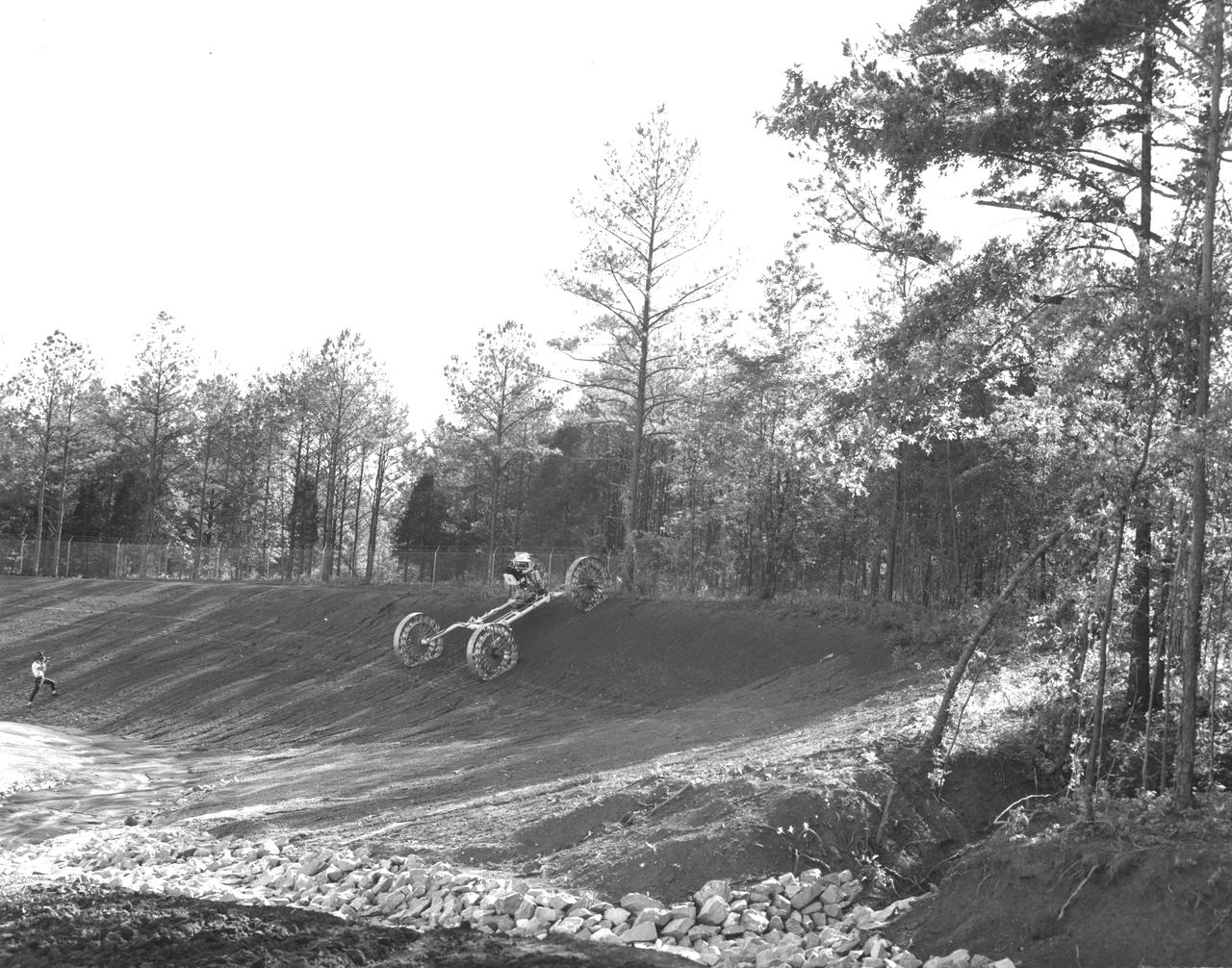An engineer demonstrates a Mobility Test Article (MTA) at NASA’s Marshall Space Flight Center (MSFC). This unit, weighing 1/6th as much as an actual vehicle, was built by the Bendix Corporation and was one of the concepts of a possible Lunar Roving Vehicle (LRV). The data provided by the MTA helped in designing the LRV, developed under the direction of MSFC. The LRV was designed to allow Apollo astronauts a greater range of mobility during lunar exploration missions. 