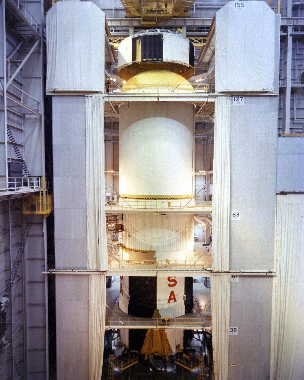 This picture is a view of stacking the major components of the S-IC (first) stage of the Saturn V vehicle at the Boeing vertical assembly building at the Michoud Assembly Facility (MAF). The view shows the S-IC forward skirt being lowered onto the liquid oxygen (LOX) tank. The Saturn IB and Saturn V first stages were manufactured at the MAF located 24 kilometers (approximately 15 miles) east of downtown New Orleans, Louisiana. The prime contractors, Chrysler and Boeing, jointly occupied the MAF. The basic manufacturing building boasted 43 acres under one roof. By 1964, NASA added a separate engineering and office building, vertical assembly building, and test stage building. By 1966, other changes to the site included enlarged barge facilities and other miscellaneous support buildings. 