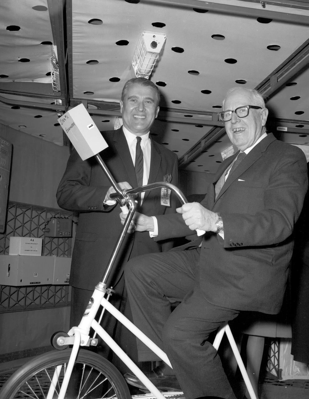 During a visit to the Marshall Space Flight Center (MSFC), the Congressional  House Committee on Science and Astronautics toured the S-IVB workshop. Pictured here are MSFC’s Dr. Wernher von Braun (standing) and Congressman Miller, Democratic representative of California (sitting on the ergometer bicycle) inside the workshop.