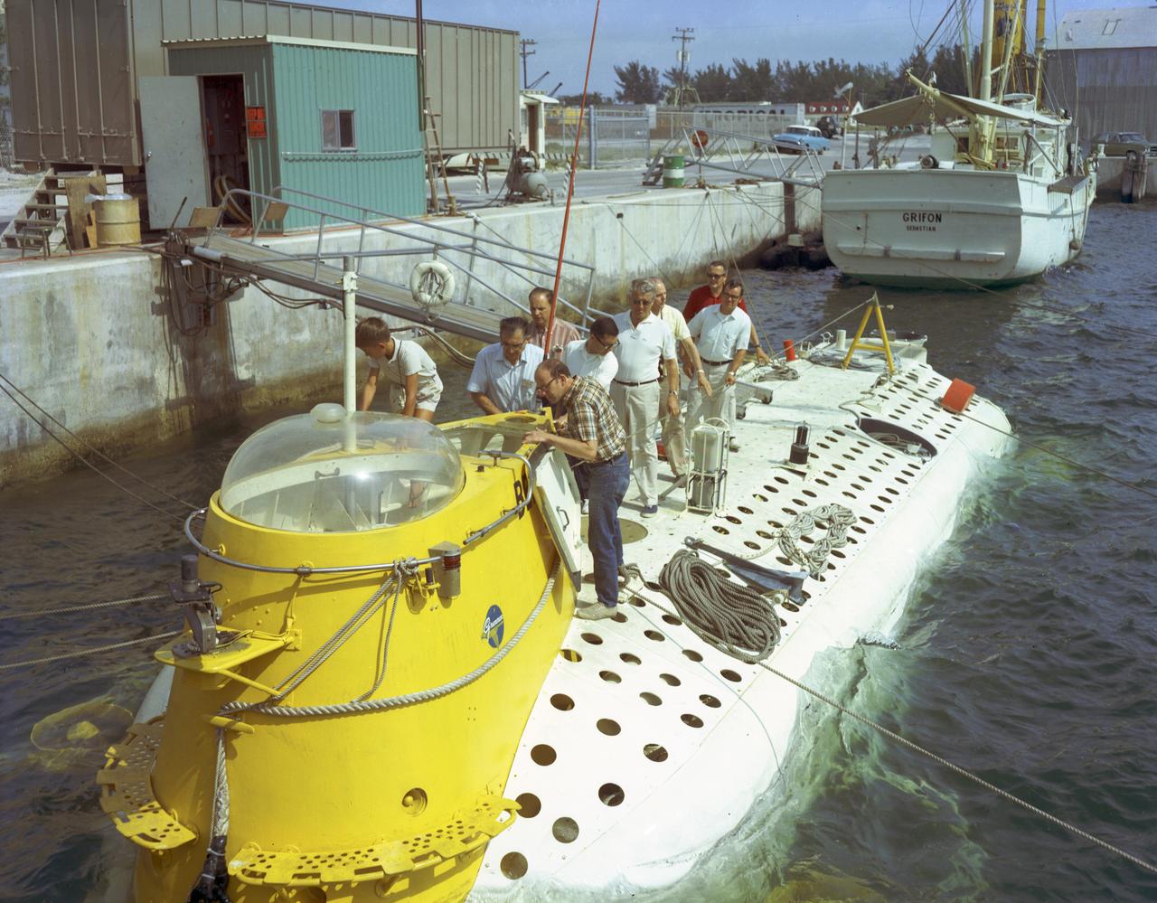 This photograph depicts Dr. von Braun (fourth from far right) and other NASA officials surveying the deep-sea research submarine "Ben Franklin." Named for American patriot and inventor Ben Franklin, who discovered the Gulf Steam, the 50-foot Ben Franklin was built between 1966 and 1968 in Switzerland for deep-ocean explorer Jacques Piccard and the Grumman Aircraft Engineering Corporation. The submersible made a famous 30-day drift dive off the East Coast of the United States and Canada in 1969 mapping the Gulf Stream's currents and sea life, and also made space exploration history by studying the behavior of aquanauts in a sealed, self-contained, self-sufficient capsule for NASA. On July 14, 1969, the Ben Franklin was towed to the high-velocity center of the Stream off the coast of Palm Beach, Florida. With a NASA observer on board, the sub descended to 1,000 feet off of Riviera Beach, Florida and drifted 1,400 miles north with the current for more than four weeks, reemerging near Maine. During the course of the dive, NASA conducted exhaustive analyses of virtually every aspect of onboard life. They measured sleep quality and patterns, sense of humor and behavioral shifts, physical reflexes, and the effects of a long-term routine on the crew. The submarine's record-shattering dive influenced the design of Apollo and Skylab missions and continued to guide NASA scientists as they devised future marned space-flight missions.