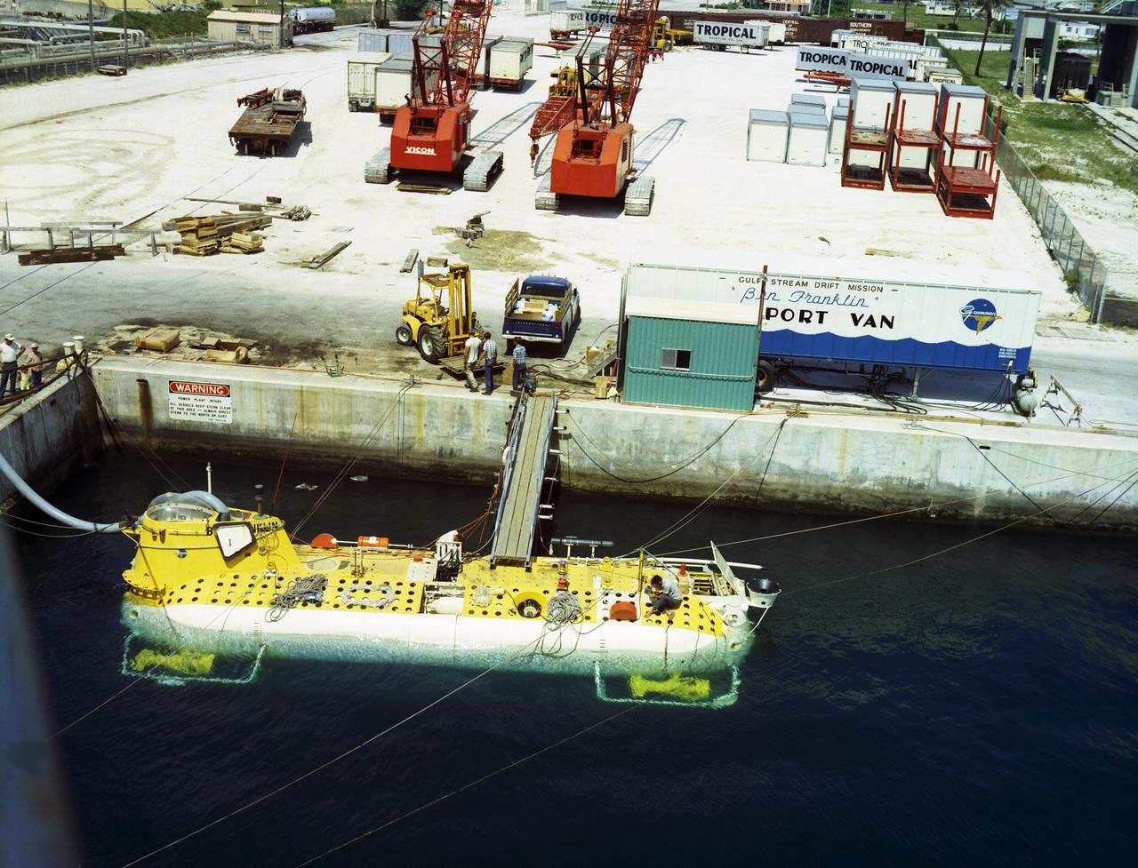 This is an aerial view of the deep-sea research submarine "Ben Franklin" at dock. Named for American patriot and inventor Ben Franklin, who discovered the Gulf Steam, the 50-foot Ben Franklin was built between 1966 and 1968 in Switzerland for deep-ocean explorer Jacques Piccard and the Grumman Aircraft Engineering Corporation. The submersible made a famous 30-day drift dive off the East Coast of the United States and Canada in 1969 mapping the Gulf Stream's currents and sea life, and also made space exploration history by studying the behavior of aquanauts in a sealed, self-contained, self-sufficient capsule for NASA. On July 14, 1969, the Ben Franklin was towed to the high-velocity center of the Stream off the coast of Palm Beach, Florida. With a NASA observer on board, the sub descended to 1,000 feet off of Riviera Beach, Florida and drifted 1,400 miles north with the current for more than four weeks, reemerging near Maine. During the course of the dive, NASA conducted exhaustive analyses of virtually every aspect of onboard life. They measured sleep quality and patterns, sense of humor and behavioral shifts, physical reflexes, and the effects of a long-term routine on the crew. The submarine's record-shattering dive influenced the design of Apollo and Skylab missions and continued to guide NASA scientists as they devised future marned space-flight missions.