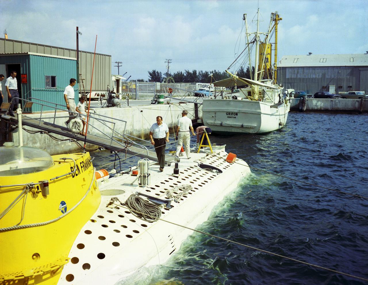 This photograph depicts Dr. von Braun (at right, showing his back) and other NASA officials surveying the deep-sea research submarine "Ben Franklin." Named for American patriot and inventor Ben Franklin, who discovered the Gulf Steam, the 50-foot Ben Franklin was built between 1966 and 1968 in Switzerland for deep-ocean explorer Jacques Piccard and the Grumman Aircraft Engineering Corporation. The submersible made a famous 30-day drift dive off the East Coast of the United States and Canada in 1969 mapping the Gulf Stream's currents and sea life, and also made space exploration history by studying the behavior of aquanauts in a sealed, self-contained, self-sufficient capsule for NASA. On July 14, 1969, the Ben Franklin was towed to the high-velocity center of the Stream off the coast of Palm Beach, Florida. With a NASA observer on board, the sub descended to 1,000 feet off of Riviera Beach, Florida and drifted 1,400 miles north with the current for more than four weeks, reemerging near Maine. During the course of the dive, NASA conducted exhaustive analyses of virtually every aspect of onboard life. They measured sleep quality and patterns, sense of humor and behavioral shifts, physical reflexes, and the effects of a long-term routine on the crew. The submarine's record-shattering dive influenced the design of Apollo and Skylab missions and continued to guide NASA scientists as they devised future marned space-flight missions.
