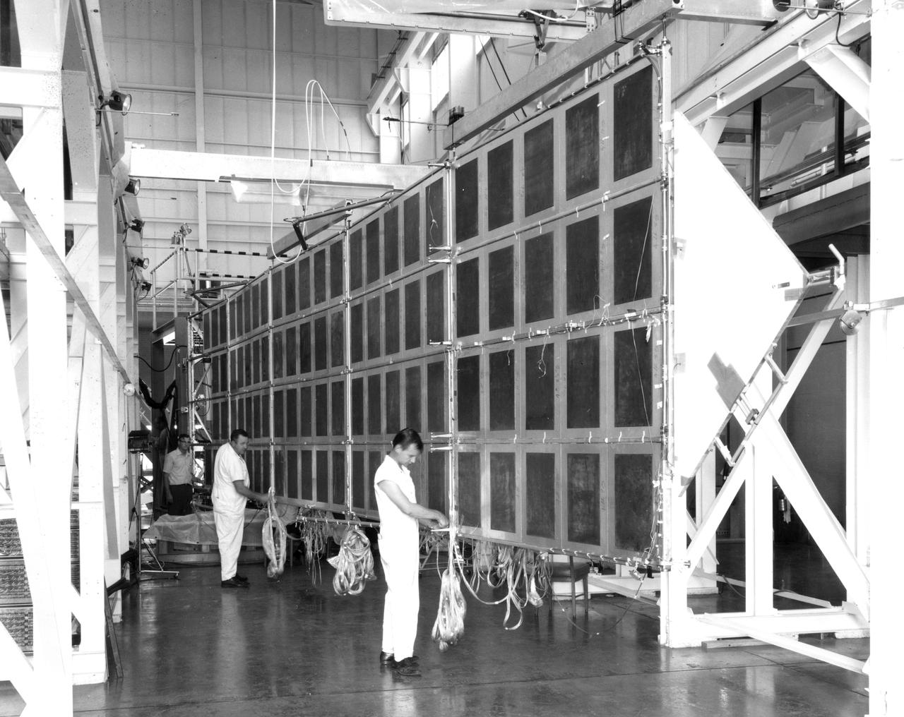 Technicians at NASA’s Marshall Space Flight Center check the wiring on a mechanical test article of the Apollo Telescope Mount (ATM) solar array.  Four such arrays were joined in a cross to provide electric power for the ATM in Earth orbit.  The deployment mechanism for extending the wing to the fully open position had just been tested when this photograph was taken.  The array was suspended from beams riding on air bearings to closely simulate the weightless conditions under which it would be deployed in space.  The wings are folded against the sides of the ATM for launch and are deployed by a scissors mechanism in Earth’s orbit.