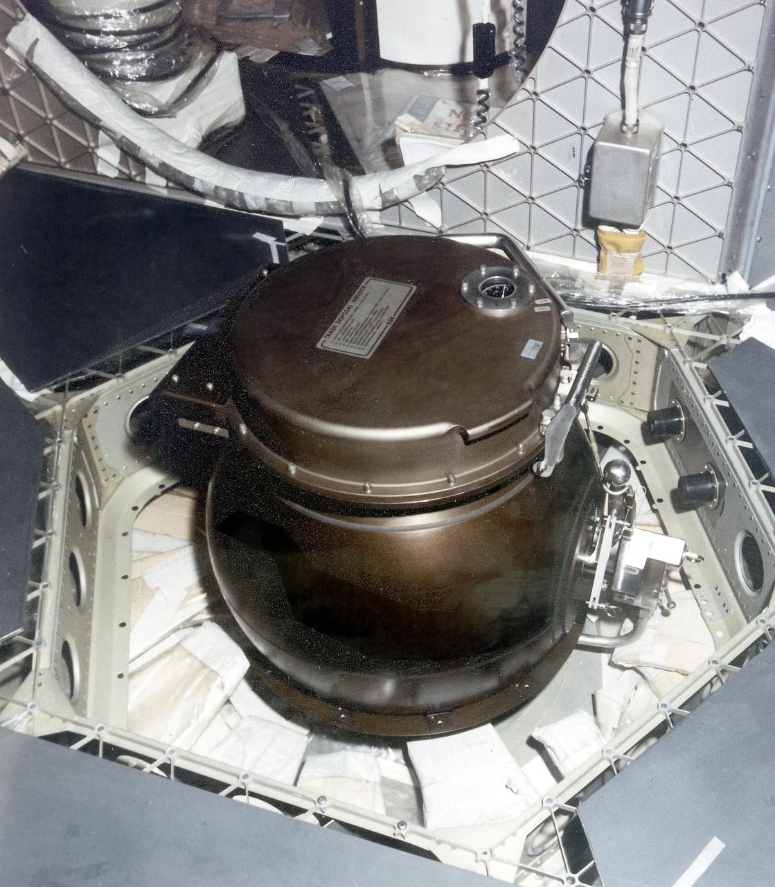 This is a close-up photograph of the Orbital Workshop (OWS) trash disposal airlock located on the floor of the lower level of the OWS. It provided a means of passing trash from the pressurized habitable area into the unpressurized waste tank. The crewman opened a valve which brought the airlock to the same pressure as that within the workshop. He then opened the lid, placed the bagged trash inside, closed the lid and locked it. By turning the valve handle, he reduced the pressure within the airlock until it reached the vacuum of the waste tank. The crewman then operated an ejector handle that caused a scissors-type mechanism to push the bagged trash from the airlock into the tank. 