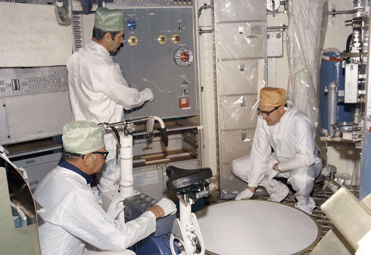 This photograph shows technicians performing a checkout of the Metabolic Analyzer (center background) and the Ergometer (foreground) in the Orbital Workshop (OWS). The shower compartment is at right. The Ergometer (Skylab Experiment M171) evaluated man's metabolic effectiveness and cost of work in space environment. Located in the experiment and work area of the OWS, the shower compartment was a cylindrical cloth enclosure that was folded flat when not in use. The bottom ring of the shower was fastened to the floor and contained foot restraints. The upper ring contained the shower head and hose. To use the shower, the astronaut filled a pressurized portable bottle with heated water and attached the bottle to the ceiling. A flexible hose cornected the water bottle to a handheld shower head. The astronaut pulled the cylindrical shower wall up into position and bathed, using liquid soap. Both soap and water were carefully rationed, having been premeasured for economical use. 