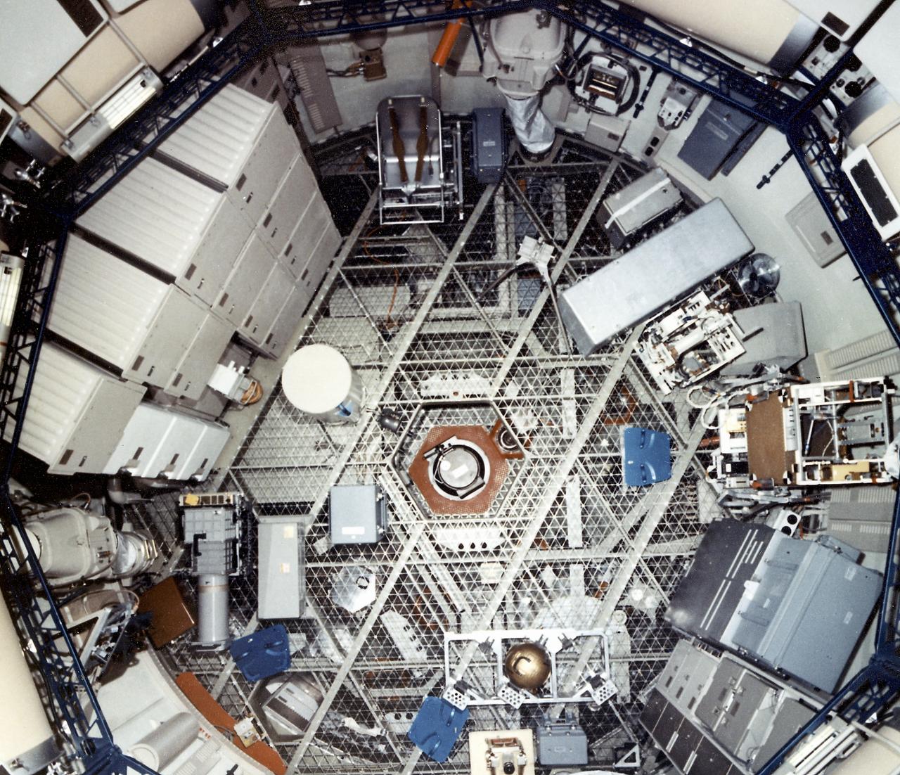 This photograph is an interior view of the Orbital Workshop (OWS) upper level looking from the airlock hatch, showing the octagonal opening that separated the workshop's two levels. The trash airlock can be seen at center. The lower level of the OWS provided crew accommodations for sleeping, food preparation and consumption, hygiene, waste processing and disposal, and performance of certain experiments. The upper level consisted of a large work area and housed water storage tanks, a food freezer, storage vaults for film, scientific airlocks, mobility and stability experiment equipment, and other experimental equipment.