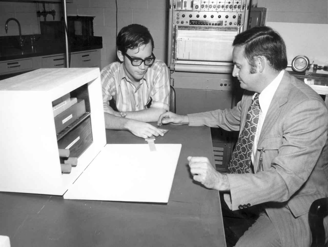San Antonio, Texas high school student, Terry C. Quist (left), and Dr. Raymond Gause of the Marshall Space Flight Center (MSFC), discuss the student’s experiment to be performed aboard the Skylab the following year. His experiment, “Earth Orbital Neutron Analysis”  required detectors such as the one he is examining in this photo.  The detector was to be attached to a water tank in Skylab. Neutrons striking the detectors left traces that were brought out by a chemical etching process after the Skylab mission. Quist’s experiment seeked to record neutron hits, count them, and determine their direction. This information was to help determine the source of neutrons in the solar system.  Quist was among 25 winners of a contest in which some 3,500 high school students proposed experiments for the following year’s Skylab mission. The nationwide scientific competition was sponsored by the National Science Teachers Association and the National Aeronautics and Space Administration (NASA). The winning students, along with their parents and sponsor teachers, visited MSFC two months earlier where they met with scientists and engineers, participated in design reviews for their experiments, and toured MSFC facilities. Of the 25 students, 6 did not see their experiments conducted on Skylab because the experiments were not compatible with Skylab hardware and timelines. Of the 19 remaining, 11 experiments required the manufacture of additional equipment. The equipment for the experiments was manufactured at MSFC.