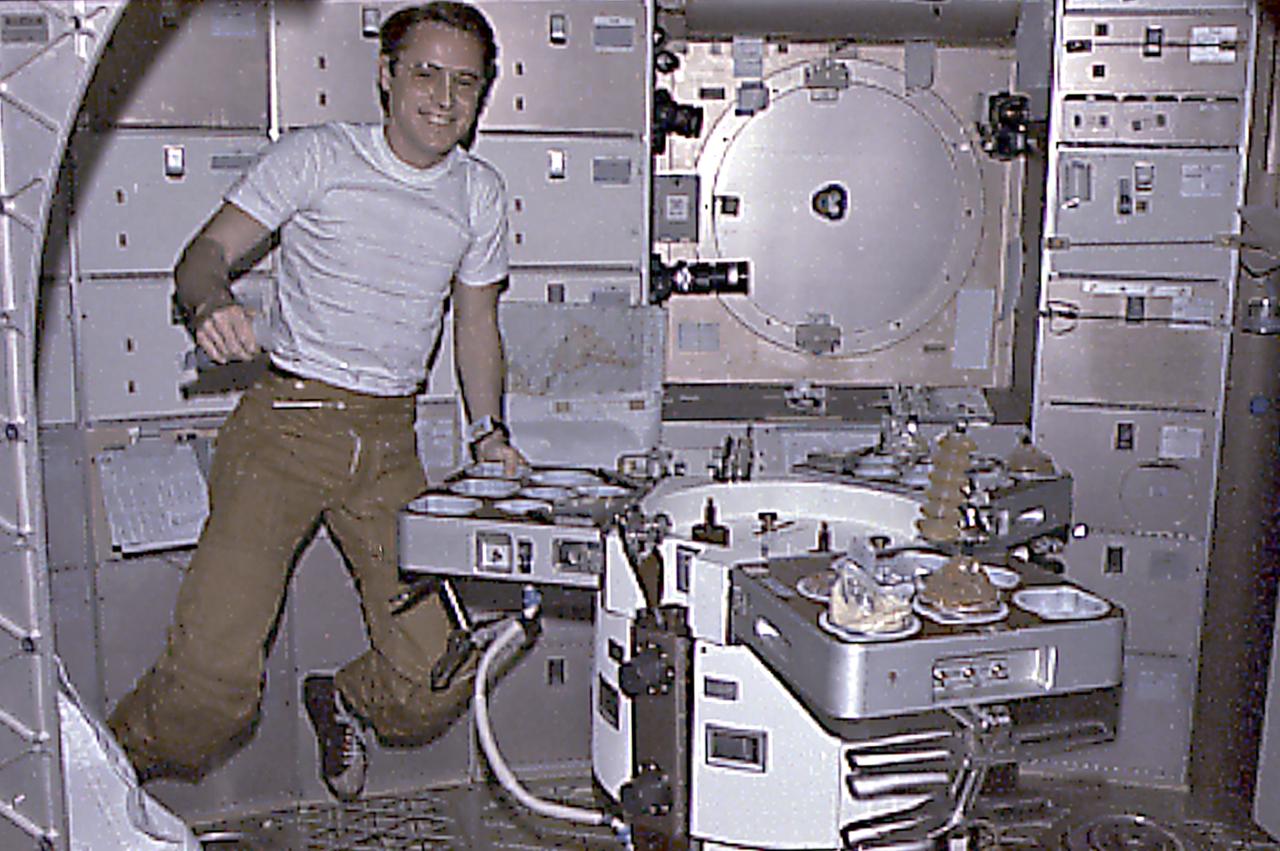 This Skylab-4 mission onboard photograph shows Astronaut Ed Gibson getting ready to prepare his meal in the crew wardroom. The tray contained heating elements for preparing the individual food packets. The food on Skylab was a great improvement over that on earlier spaceflights. It was no longer necessary to squeeze liquified food from plastic tubes. Skylab's kitchen was so equipped that each crewman could select his own menu and prepare it to his own taste.