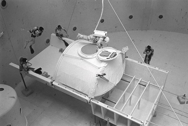 Once the United States' space program had progressed from Earth's orbit into outerspace, the prospect of building and maintaining a permanent presence in space was realized. To accomplish this feat, NASA launched a temporary workstation, Skylab, to discover the effects of low gravity and weightlessness on the human body, and also to develop tools and equipment that would be needed in the future to build and maintain a more permanent space station. The structures, techniques, and work schedules had to be carefully designed to fit this unique construction site. The components had to be lightweight for transport into orbit, yet durable. The station also had to be made with removable parts for easy servicing and repairs by astronauts. All of the tools necessary for service and repairs had to be designed for easy manipulation by a suited astronaut. Construction methods had to be efficient due to the limited time the astronauts could remain outside their controlled environment. In lieu of all the specific needs for this project, an environment on Earth had to be developed that could simulate a low gravity atmosphere. A Neutral Buoyancy Simulator (NBS) was constructed by NASA Marshall Space Flight Center (MSFC) in 1968. Since then, NASA scientists have used this facility to understand how humans work best in low gravity and also provide information about the different kinds of structures that can be built. With the help of the NBS, building a space station became more of a reality. Pictured is Astronaut Paul Weitz training on a mock-up of Spacelab's airlock-hatch cover. Training was also done on the use of foot restraints which had recently been developed to help astronauts maintain their positions during space walks rather than having their feet float out from underneath them while they tried to perform maintenance and repair operations. Every aspect of every space mission was researched and demonstrated in the NBS. Using the airlock hatch cover and foot restraints were just a small example of the preparation that went into each mission.