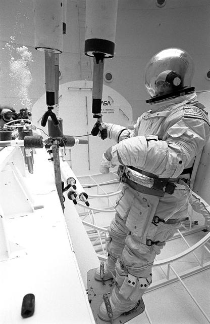 Once the United States' space program had progressed from Earth's orbit into outerspace, the prospect of building and maintaining a permanent presence in space was realized. To accomplish this feat, NASA launched a temporary workstation, Skylab, to discover the effects of low gravity and weightlessness on the human body, and also to develop tools and equipment that would be needed in the future to build and maintain a more permanent space station. The structures, techniques, and work schedules had to be carefully designed to fit this unique construction site. The components had to be lightweight for transport into orbit, yet durable. The station also had to be made with removable parts for easy servicing and repairs by astronauts. All of the tools necessary for service and repairs had to be designed for easy manipulation by a suited astronaut. Construction methods had to be efficient due to the limited time the astronauts could remain outside their controlled environment. In lieu of all the specific needs for this project, an environment on Earth had to be developed that could simulate a low gravity atmosphere. A Neutral Buoyancy Simulator (NBS) was constructed by NASA's Marshall Space Flight Center (MSFC) in 1968. Since then, NASA scientists have used this facility to understand how humans work best in low gravity and also provide information about the different kinds of structures that can be built. Pictured is a Massachusetts Institute of Technology (MIT) student working in a spacesuit on the Experimental Assembly of Structures in Extravehicular Activity (EASE) project which was developed as a joint effort between MFSC and MIT. The EASE experiment required that crew members assemble small components to form larger components, working from the payload bay of the space shuttle. The MIT student in this photo is assembling two six-beam tetrahedrons.