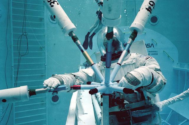 Once the United States' space program had progressed from Earth's orbit into outerspace, the prospect of building and maintaining a permanent presence in space was realized. To accomplish this feat, NASA launched a temporary workstation, Skylab, to discover the effects of low gravity and weightlessness on the human body, and also to develop tools and equipment that would be needed in the future to build and maintain a more permanent space station. The structures, techniques, and work schedules had to be carefully designed to fit this unique construction site. The components had to be lightweight for transport into orbit, yet durable. The station also had to be made with removable parts for easy servicing and repairs by astronauts. All of the tools necessary for service and repairs had to be designed for easy manipulation by a suited astronaut. And construction methods had to be efficient due to limited time the astronauts could remain outside their controlled environment. In lieu of all the specific needs for this project, an environment on Earth had to be developed that could simulate a low gravity atmosphere. A Neutral Buoyancy Simulator (NBS) was constructed by NASA Marshall Space Flight Center (MSFC) in 1968. Since then, NASA scientists have used this facility to understand how humans work best in low gravity and also provide information about the different kinds of structures that can be built. Pictured is a Massachusetts Institute of Technology (MIT) student working in a spacesuit on the Experimental Assembly of Structures in Extravehicular Activity (EASE) project which was developed as a joint effort between MFSC and MIT. The EASE experiment required that crew members assemble small components to form larger components, working from the payload bay of the space shuttle.