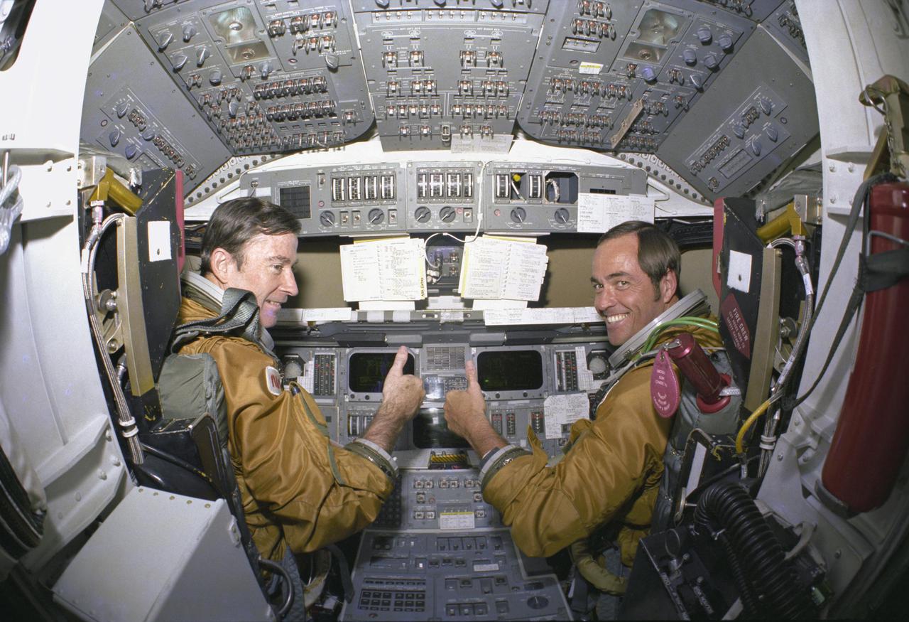 KENNEDY SPACE CENTER, FLA.  - Prime crew astronauts for the first space shuttle mission, Commander John Young and Pilot Robert Crippen, take a break from their intensive training schedule to pose for pictures in the flight deck of the orbiter Columbia.  The Space Shuttle is controlled and most payloads are handled from the flight deck.  Looking forward, the ship’s commander is seated on the left and the pilot on the right.  Shown are the TV-like displays, and duplicate sets of conventional-looking hand controllers, pedals, levers and switches with which either astronaut can fly the craft alone.  The two-man crew are wearing their ejection escape suits which will be worn only during the orbital flight test program and then only during the launch and landing phases of the mission.