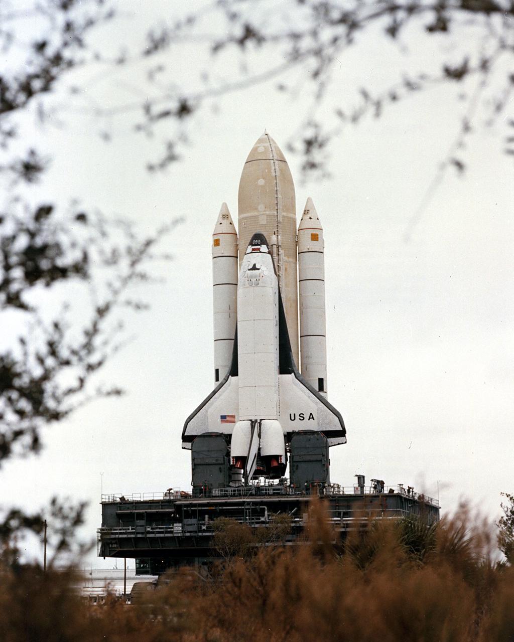 KENNEDY SPACE CENTER, FLA. - STS-1 stands framed in vegetation lining the superhighay-wide crawlerway linking the Vehicle Assembly Building with the twin pads of Launch Complex 39. The first Space Shuttle assembly emerged from the VAB shortly after 8 a.m. today for the 3.5-mile journey to Complex 39's Pad A. The Shuttle was on the hardstand at Pad A approximately seven and a half hours after rollout began. Launch is scheduled for no eariler than March 1981.