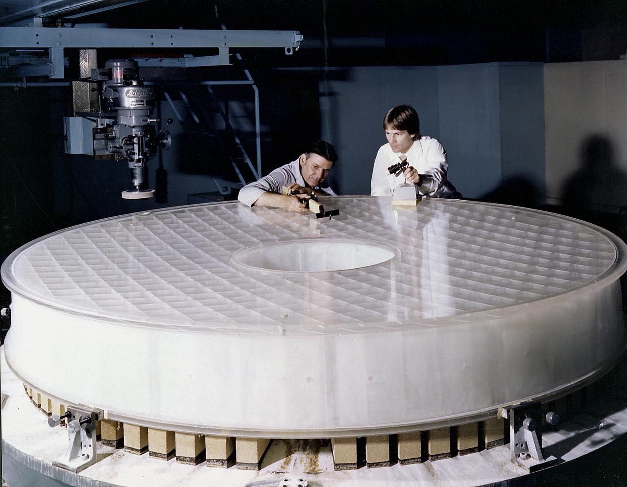 This photograph shows engineers inspecting the Hubble Space Telescope's (HST's) Primary Mirror at the Perkin-Elmer Corporation's large optics fabrication facility. After the 8-foot diameter mirror was ground to shape and polished, the glass surface was coated with a reflective layer of aluminum and a protective layer of magnesium fluoride, 0.1- and 0.025- micrometers thick, respectively. The purpose of the HST, the most complex and sensitive optical telescope ever made, is to study the cosmos from a low-Earth orbit. By placing the telescope in space, astronomers are able to collect data that is free of the Earth's atmosphere. The Marshall Space Flight Center had responsibility for design, development, and construction of the HST and the Perkin-Elmer Corporation, in Danbury, Cornecticut, developed the optical system and guidance sensors.