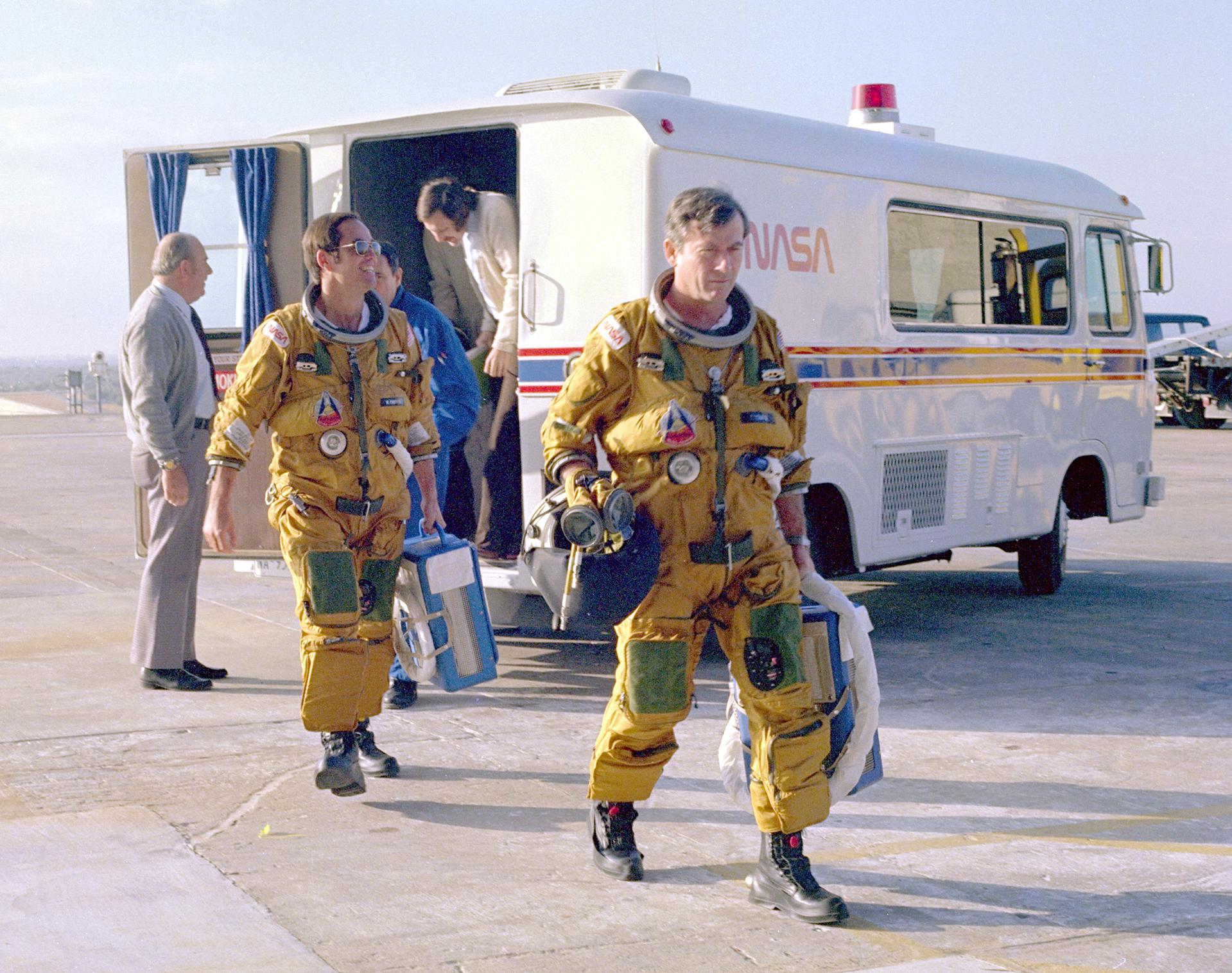 Space Shuttle prime astronaut crew members Bob Crippen and John Young in yellow flight suits walk away from a NASA van they just departed