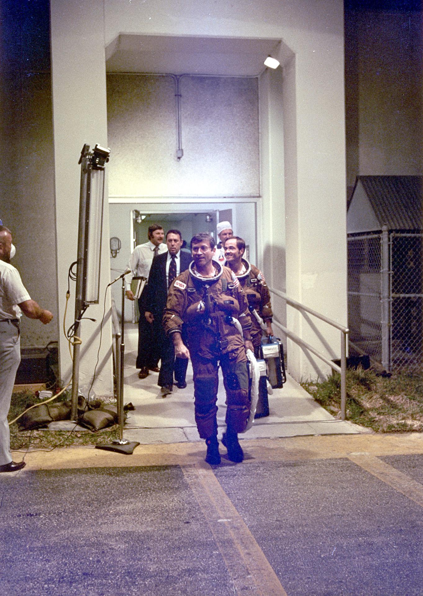 The STS-1 Prime Crew Astronauts, John Young (in front) and Bob Crippen, are followed by Flight Operations Director George Abbey, crew trainer Richard Nygram, and a space suit technician as they depart KSC’s Operations and Checkout Building