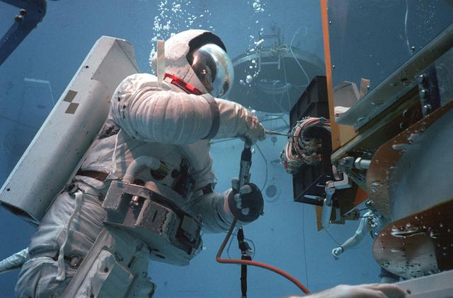 In February 1980, a satellite called Solar Maximum Mission Spacecraft, or Solar Max, was launched into Earth's orbit. Its primary objective was to provide a detailed study of solar flares, active regions on the Sun's surface, sunspots, and other solar activities. Additionally, it was to measure the total output of radiation from the Sun. Not much was known about solar activity at that time except for a slight knowledge of solar flares. After its launch, Solar Max fulfilled everyone's expectations. However, after a year in orbit, Solar Max's Altitude Control System malfunctioned, preventing the precise pointing of instruments at the Sun. NASA scientists were disappointed at the lost data, but not altogether dismayed because Solar Max had been designed for Space Shuttle retrievability enabling the repair of the satellite. On April 6, 1984, Space Shuttle Challenger (STS-41C), Commanded by astronaut Robert L. Crippen and piloted by Francis R. Scobee, launched on a historic voyage. This voyage initiated a series of firsts for NASA; the first satellite retrieval, the first service use of a new space system called the Marned Maneuvering Unit (MMU), the first in-orbit repair, the first use of the Remote Manipulator System (RMS), and the Space Shuttle Challenger's first space flight. The mission was successful in retrieving Solar Max. Mission Specialist Dr. George D. Nelson, using the MMU, left the orbiter's cargo bay and rendezvoused with Solar Max. After attaching himself to the satellite, he awaited the orbiter to maneuver itself nearby. Using the RMS, Solar Max was captured and docked in the cargo bay while Dr. Nelson replaced the altitude control system and the coronagraph/polarimeter electronics box. After the repairs were completed, Solar Max was redeposited in orbit with the assistance of the RMS. Prior to the April 1984 launch, countless man-hours were spent preparing for this mission. The crew of Challenger spent months at Marshall Space Flight Center's (MSFC) Neutral Buoyancy Simulator (NBS) practicing retrieval maneuvers, piloting the MMU, and training on equipment so they could make the needed repairs to Solar Max. Pictured is Dr. Nelson performing a replacement task on the Solar Max mock-up in the NBS.