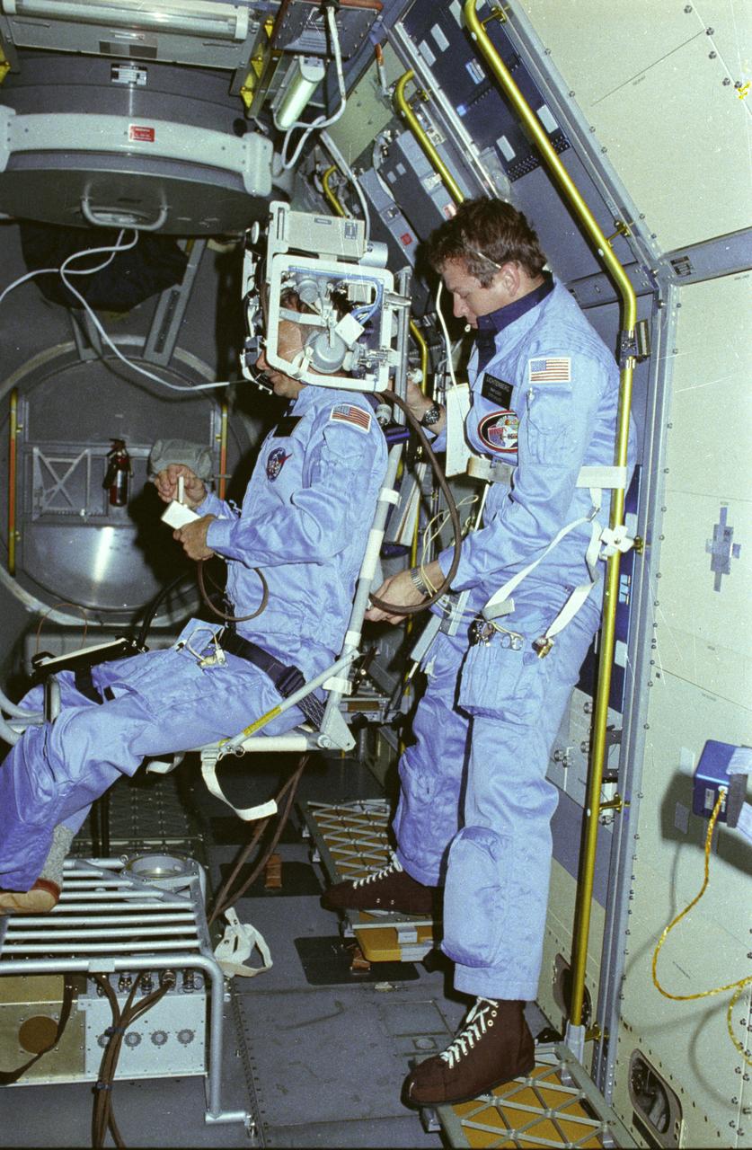 In this photograph, astronauts Owen Garriott on the body restriant system and Byron Lichtenberg prepare for a Vestibular Experiment during the Spacelab-1 mission. The Vestibular Experiments in Space were the study of the interaction among the otoliths, semicircular canals, vision, and spinal reflexes in humans. The main objective was to determine how the body, which receives redundant information for several sensory sources, interprets this information in microgravity. Another objective was to record and characterize the symptoms of space sickness experienced by crewmembers. The body restraint system was a rotating chair with a harness to hold the test subject in place. The crewmember wore an accelerometer and electrodes to record head motion and horizontal and vertical eye movement as the body rotated. The first Spacelab mission, Spacelab-1, sponsored jointly and shared equally by NASA and the European Space Agency, was a multidisciplinary mission; that is, investigations were performed in several different fields of scientific research. The overall goal of the mission was to verify Spacelab performance through a variety of scientific experiments. The Spacelab-1 was launched aboard the Space Shuttle Orbiter Columbia for the STS-9 mission on November 28, 1983. The Marshall Space Flight Center had management responsibilities for the mission.