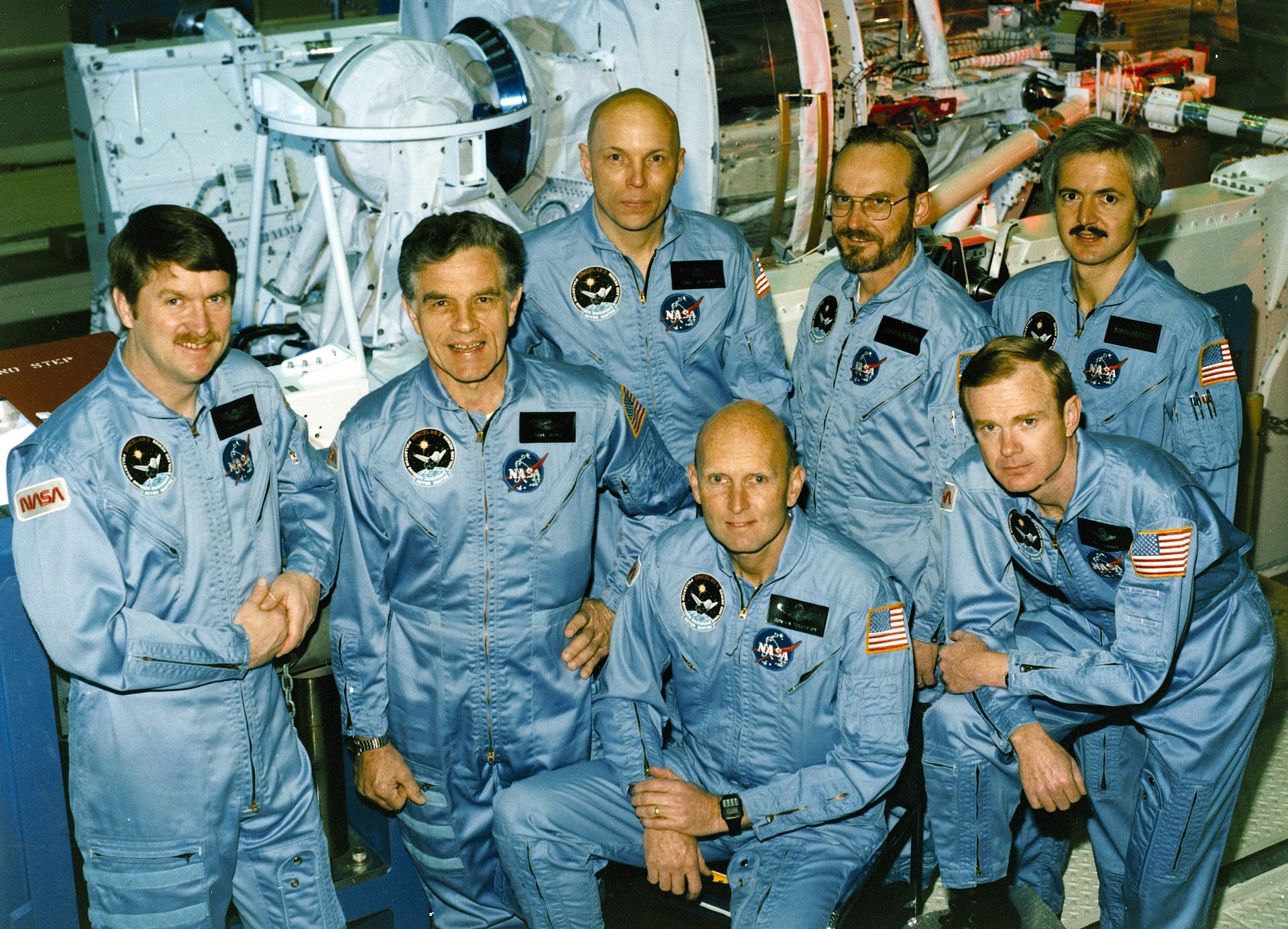 Seven men stand and smile for their group photo. They are wearing space flight suites with NASA emblems on them. 