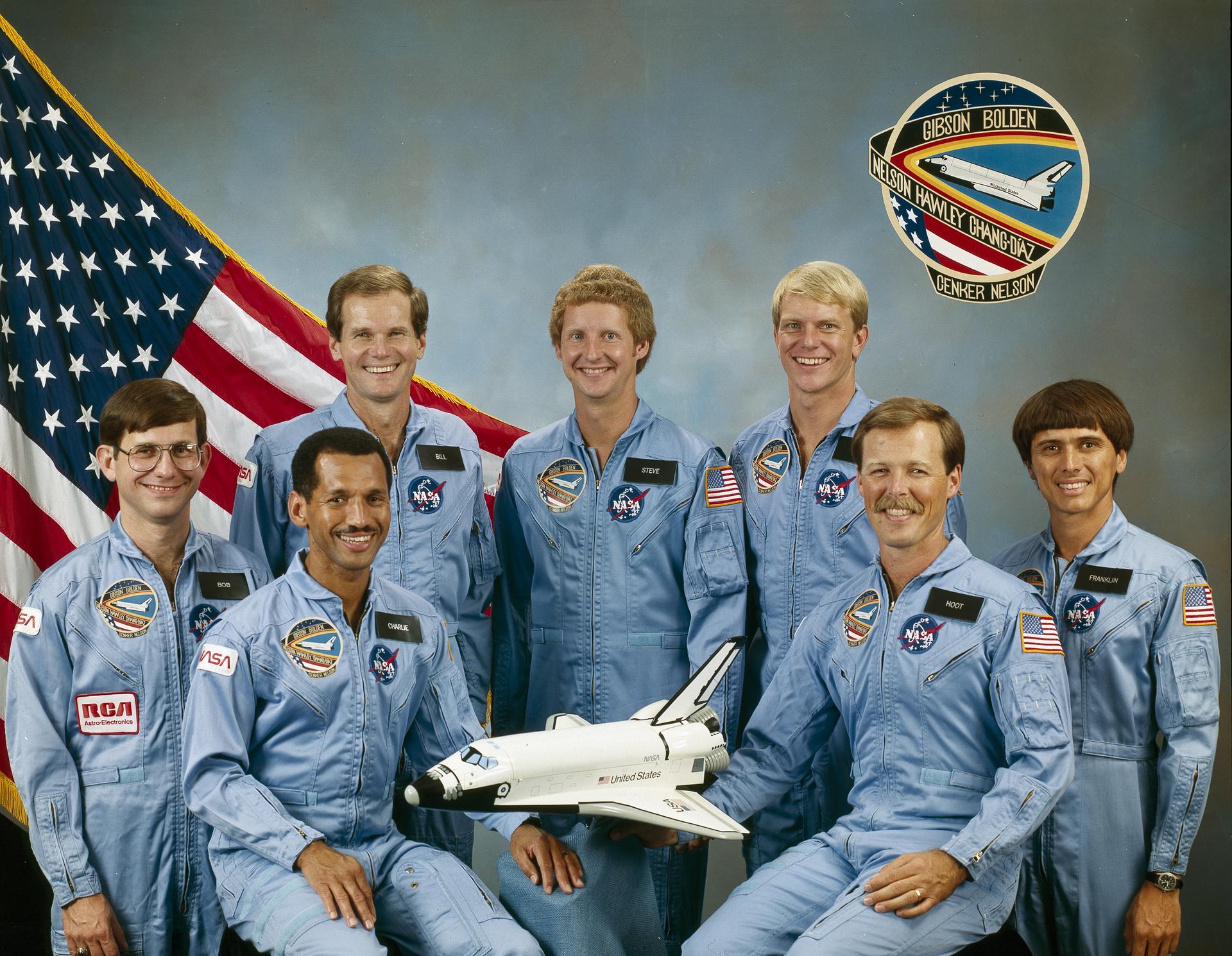 Seven astronauts pose for their crew picture. They are wearing space flight suits and there is a small model rocket ship before them.