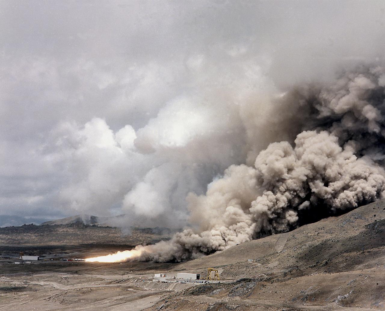 This photograph is a long shot view of a full scale solid rocket motor (SRM) for the solid rocket booster (SRB) being test fired at Morton Thiokol's Wasatch Operations in Utah. The twin boosters provide the majority of thrust for the first two minutes of flight, about 5.8 million pounds, augmenting the Shuttle's main propulsion system during liftoff. The major design drivers for the SRM's were high thrust and reuse. The desired thrust was achieved by using state-of-the-art solid propellant and by using a long cylindrical motor with a specific core design that allows the propellant to burn in a carefully controlled marner. Under the direction of the Marshall Space Flight Center, the SRM's are provided by the Morton Thiokol Corporation.
