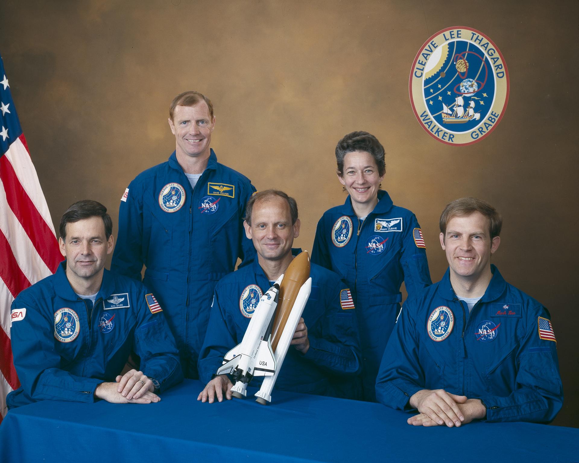 Five astronauts in blue jump suits pose with model shuttle in front of US flag.