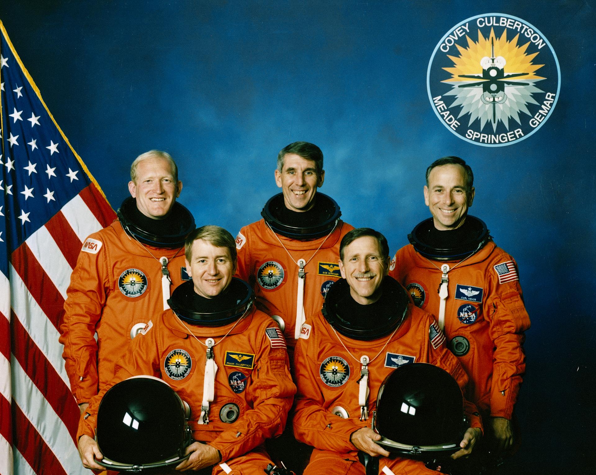 Five men in orange spacesuits pose in front of US flag