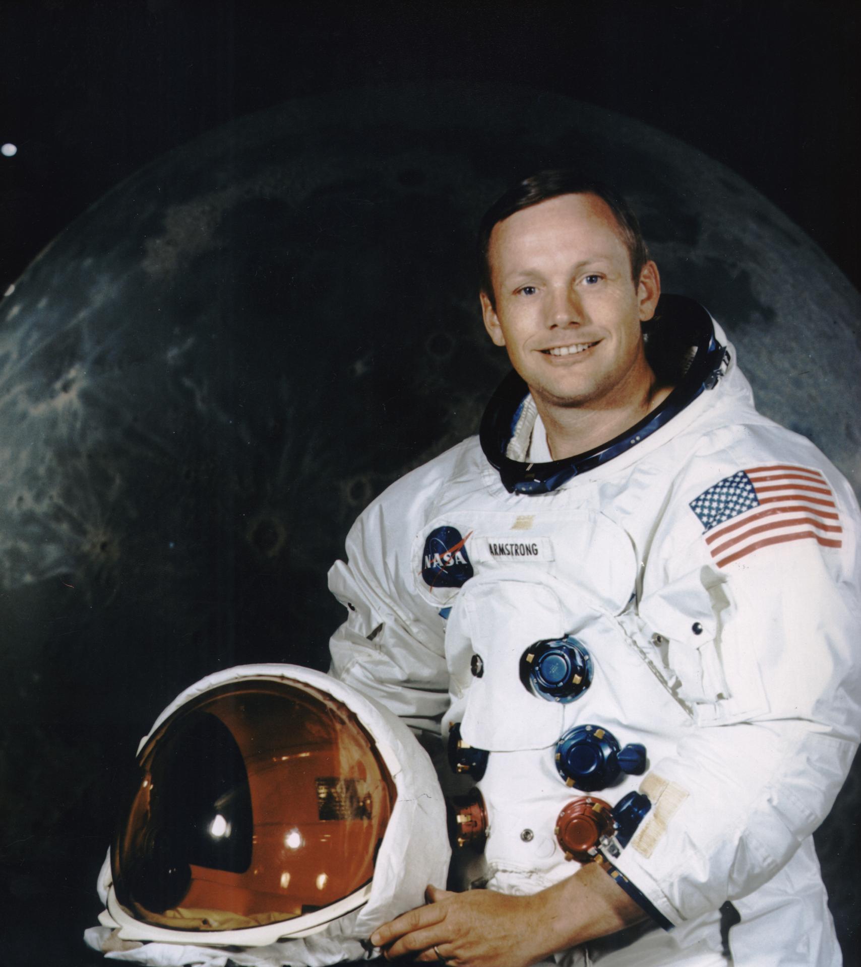 A man stands before an image of the moon, dressed in a spacesuit. His helmet is off and held before him.