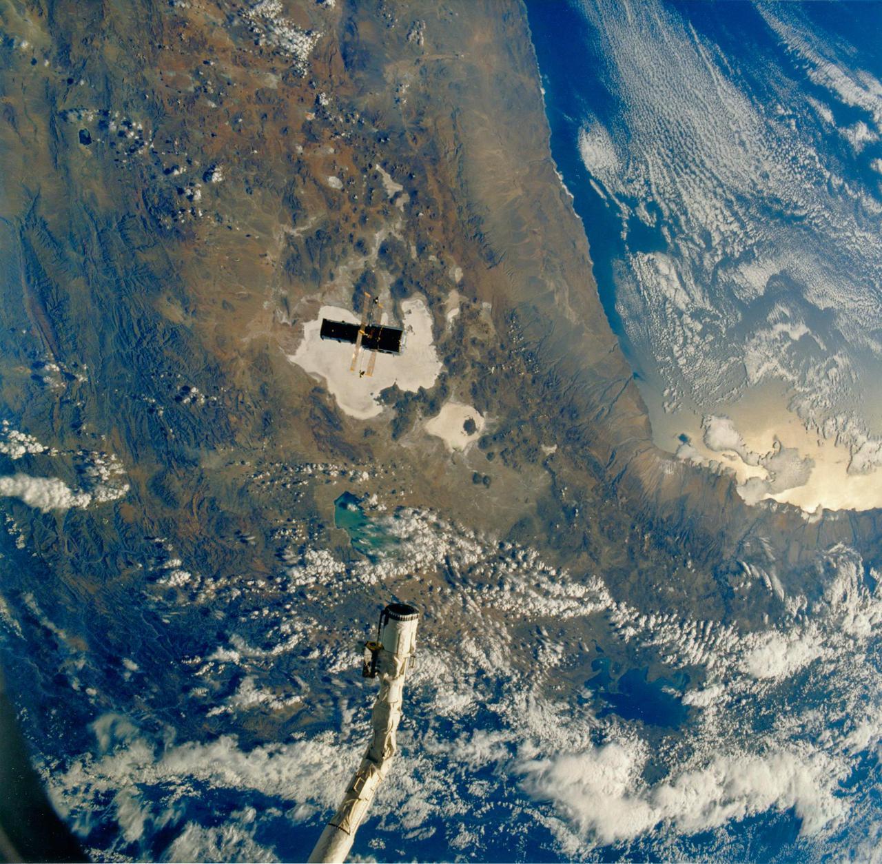 JOHNSON SPACE CENTER, HOUSTON, TEXAS -- STS-31 ONBOARD SCENE -- The huge Hubble Space Telescope (HST), backdropped against Bolivian, Chilean and Peruvian topography some 332 nautical miles away, and the Space Shuttle Discovery, represented in this scene by its remote manipulator system (RMS) arm, begin their relative separation in space.  The chalk-colored feature directly beneath HST is called Salar de Uyuni.  Lake Titicaca is in upper right, and Lake Fuope is under partial cloud cover near the RMS's end effector.  This photograph, one of the first eight STS-31 onboard frames to be released by NASA, was taken with a handheld Hasselblad camera by one of the five busy crew members in Discovery's cabin.