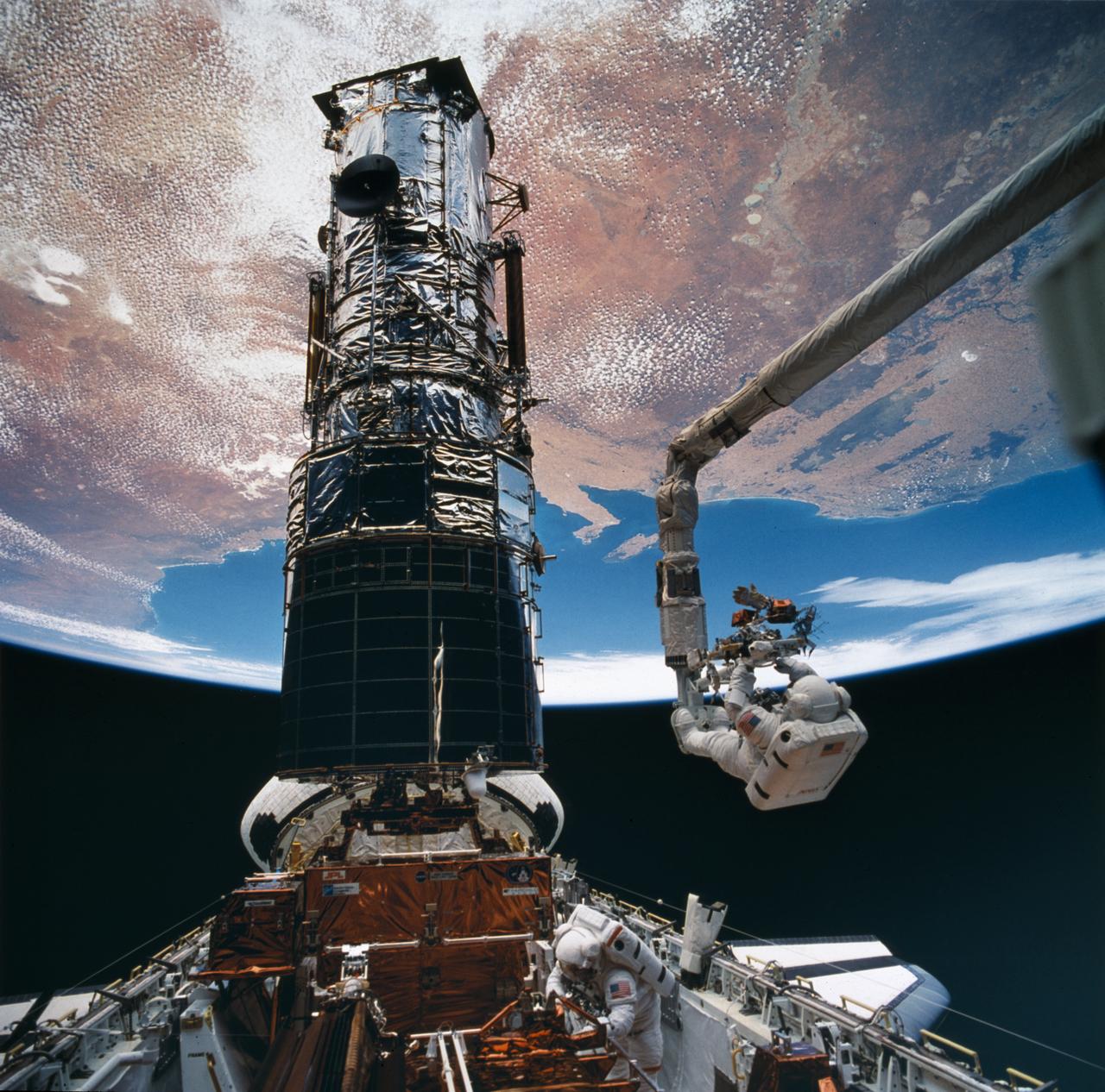 Astronaut Story Musgrave, anchored to the end of the remote manipulator arm, prepares to be elevated to the top of the towering Hubble Space Telescope (HST) to install protective covers on magnetometers. Astornaut Jeffrey Hoffman assisted Musgrave with the final servicing tasks. 