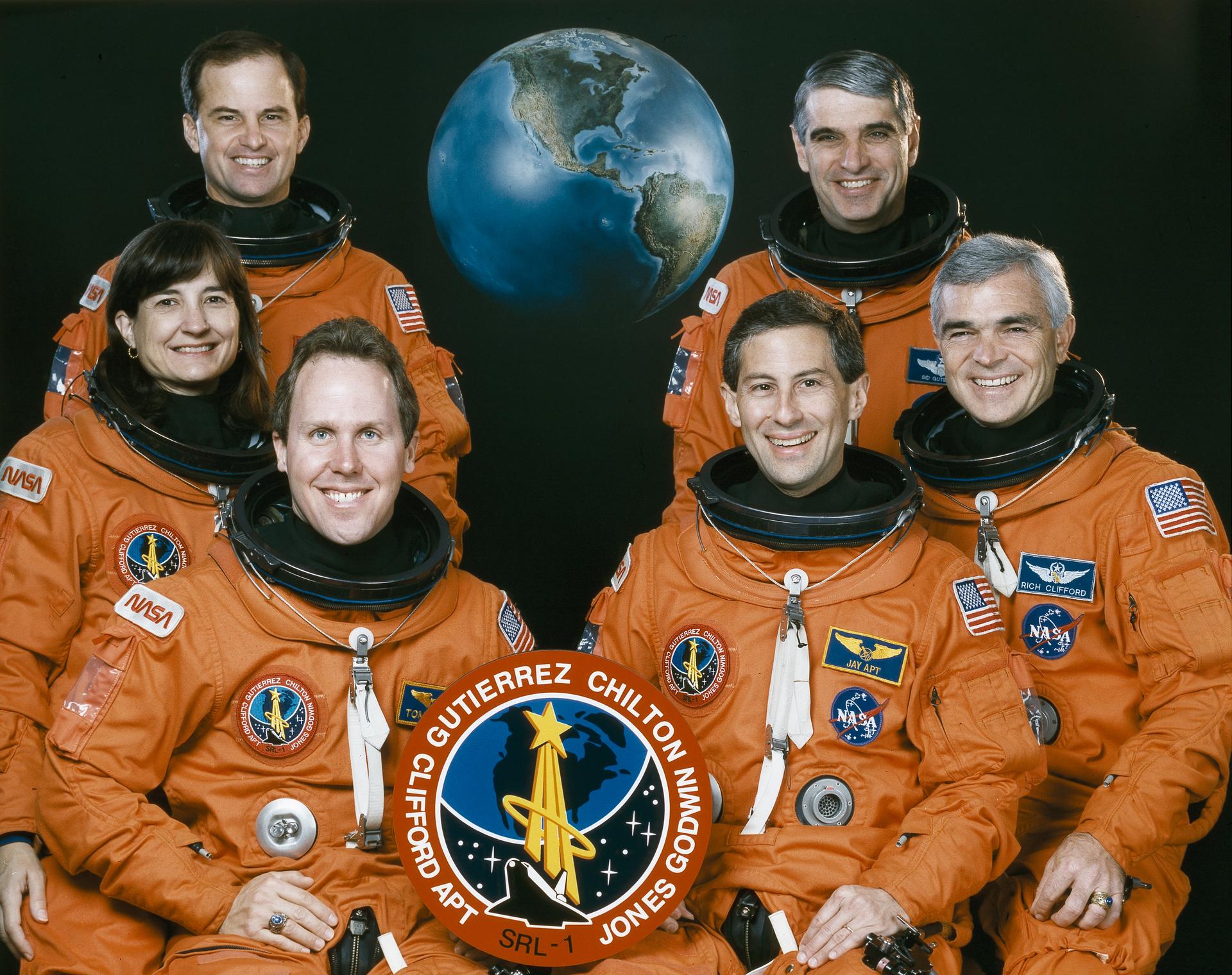 Five men and one woman in orange spacesuits pose for crew photo in front of an earth backdrop.