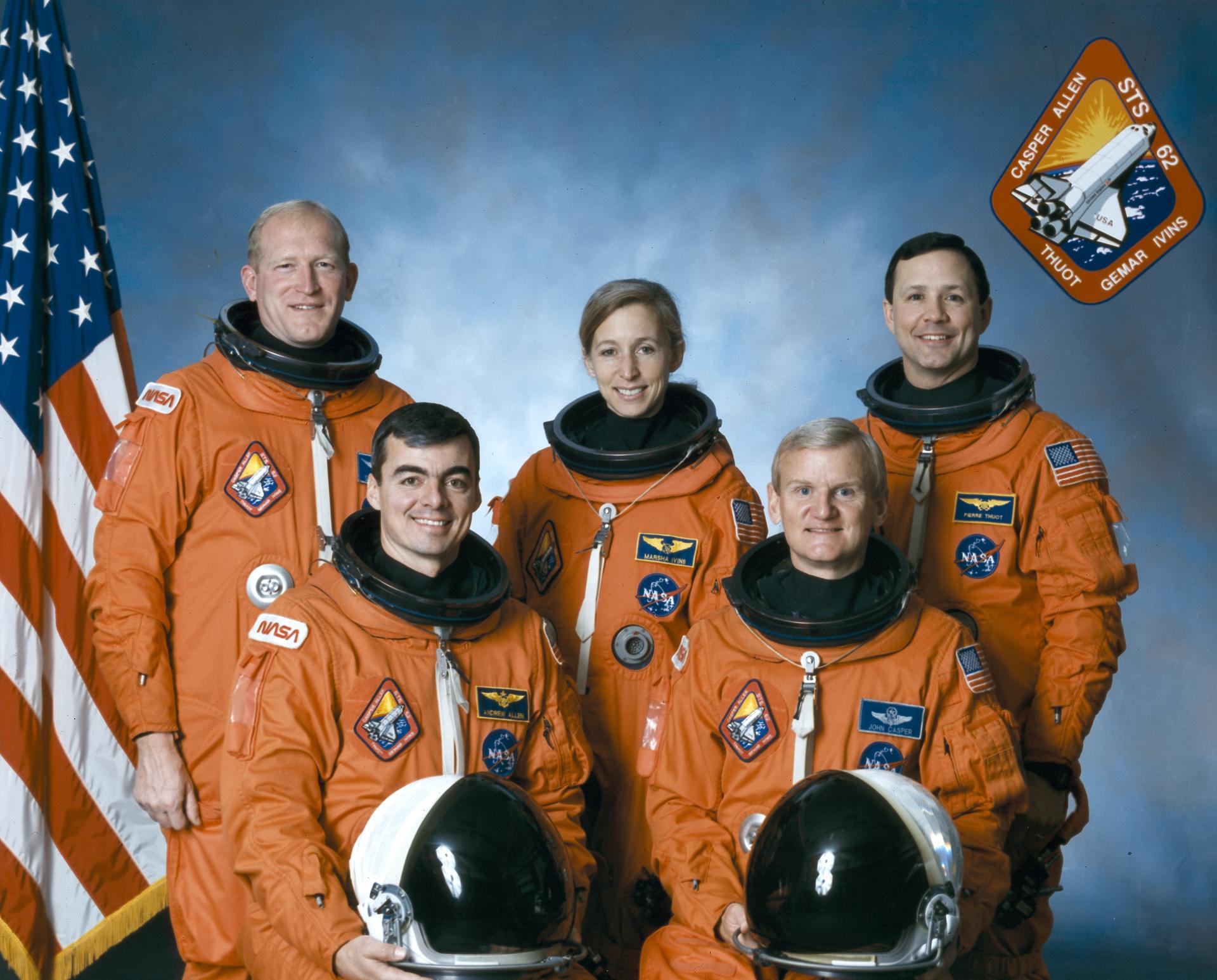 Four men and one woman in orange spacesuits pose for crew photo in front of American flag.