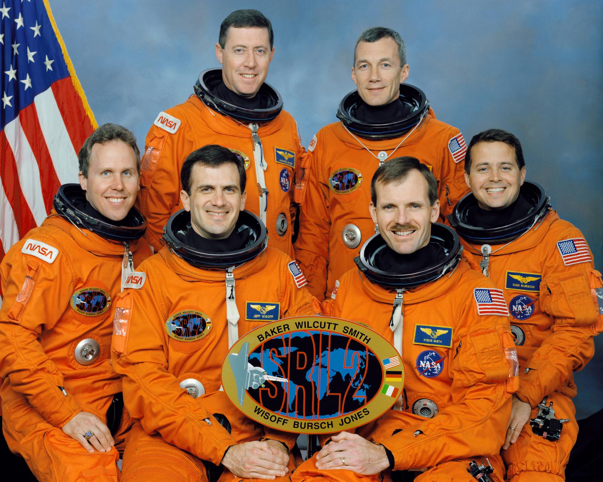 Six astronauts in orange spacesuits pose for crew photo with a BAKER WILCUTT SMITH AND WISOFF BURSCH JONES sign.