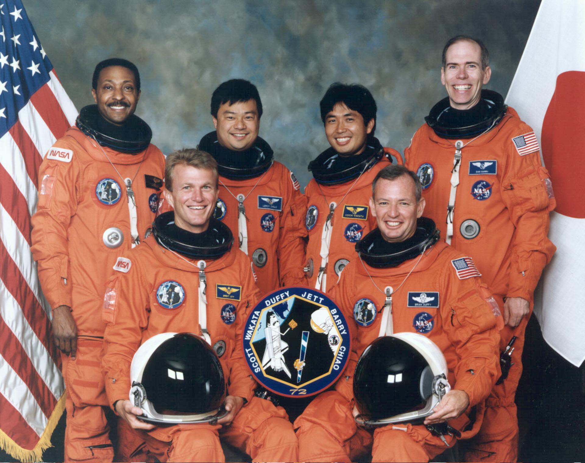 Six astronauts in orange spacesuits pose for crew photo in front of an American and Japanese flag.