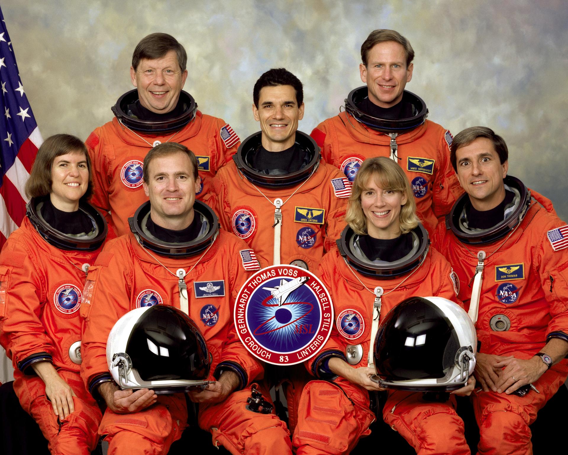 Seven astronauts pose for crew photo in orange spacesuits in front of American flag