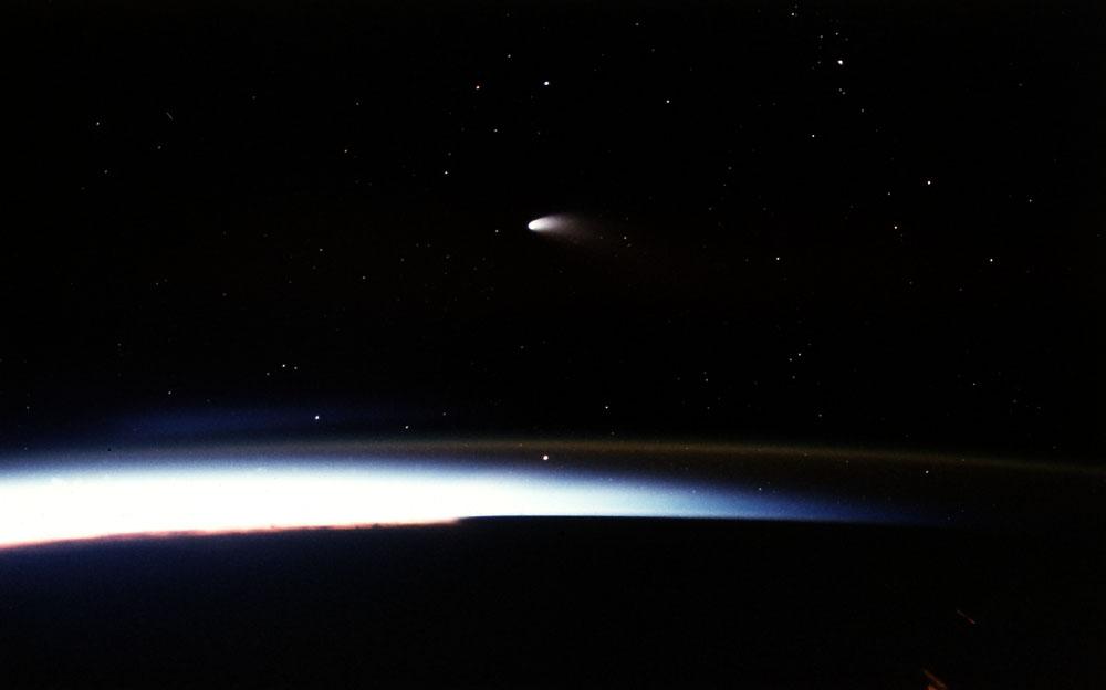This is a Microgravity Science Laboratory-1 (MLS-1) onboard STS-83 photo of the most recent comet to date, Hale-Bopp, which passed by Earth during the spring and summer of 1997. In this view, the comet is visible during sunset. The streaks and distorted lights seen in the bottom of the photo are city lights and petroleum fires.