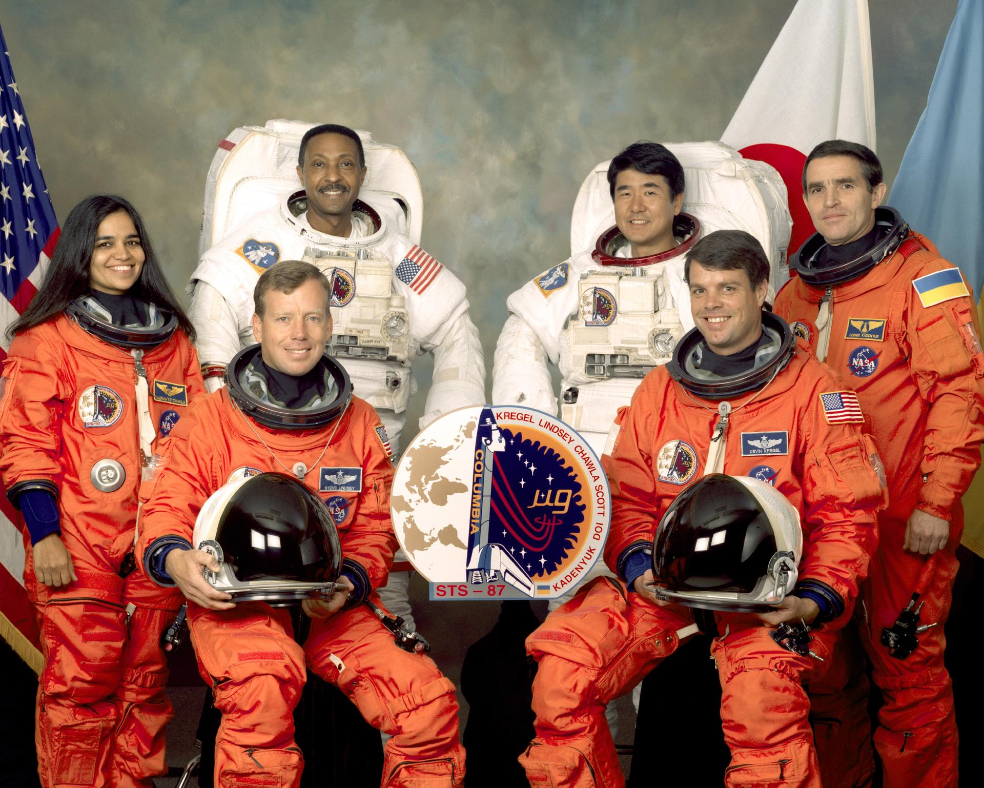 Five astronauts and a payload specialist in orange and white spacesuits pose for portrait.