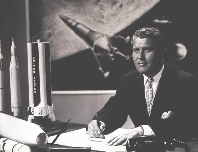 Dr. Wernher von Braun, Marshall's first Center Director (1960-1970), in his office with illustrations of rocket models behind him.