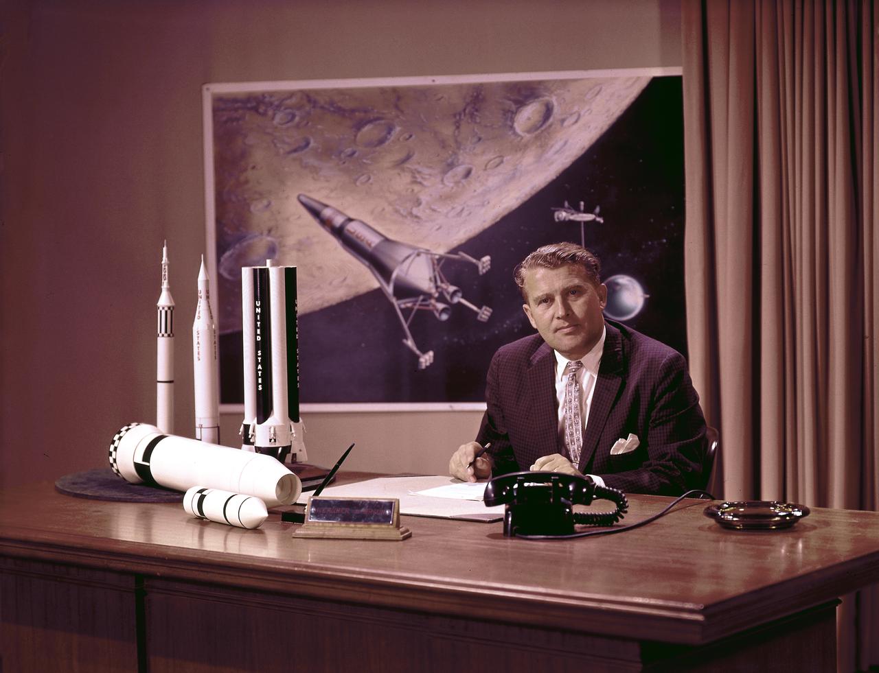 Photo of Marshall Space Flight Center (MSFC) Director Dr. Wernher von Braun at his desk with moon lander in background and rocket models on his desk. Dr. von Braun served as Marshall's first director from 1960 until his transfer to NASA Headquarters in 1970