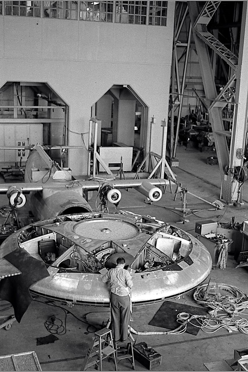 Avrocar in the shop of the 40x80 foot wind tunnel with the 4 prop tilt wing model in the back ground.