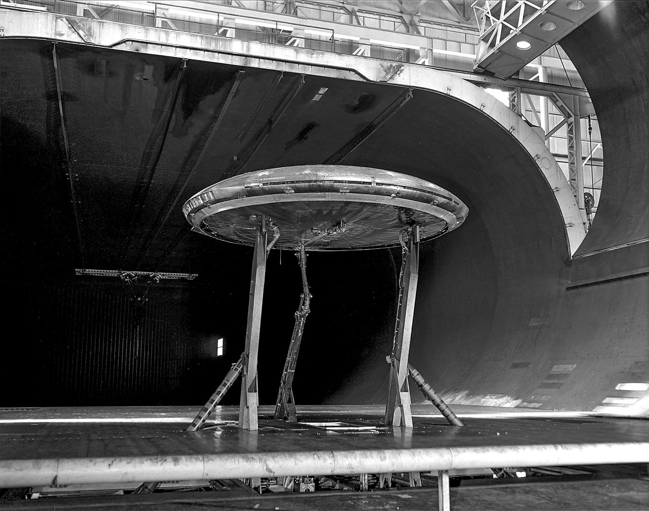 Rear view of the Avrocar with tail, mounted on variable height struts.  Overhead doors of the wind tunnel test section open. The first Avrocar, S/N 58-7055 (marked AV-7055), after tethered testing, became the "wind tunnel" test model at NASA Ames, where it remained in storage from 1961 until 1966, when it was donated to the National Air and Space Museum, in Suitland, Maryland.