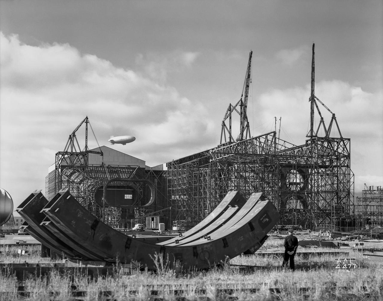 Looking south at the construction of the test section of the 40 x 80 foot wind tunnel.  4 parts of a test section bent seen in the  foreground. Airship rising in the background.