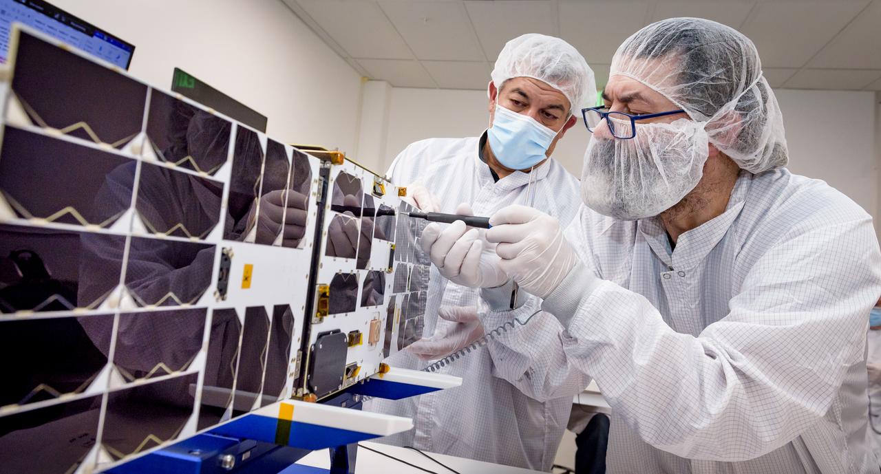 Mario Perez, back, holds the deployable solar panel as Craig Turczynski, left, secures it to the Advanced Composite Solar Sail System (ACS3) spacecraft in the Ames Integration Facility located in N213 room 104.