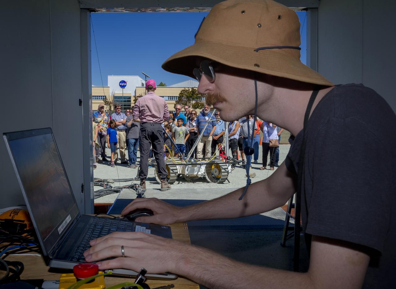 Ethan Massey, in the control booth, sends commands that control the movement of the Moon Gravity Representative Unit (MGRU3) at the Roverscape during Family Day for team members of the Volatiles Investigating Polar Exploration Rover (VIPER).  MGRU3 is a weight equivalent mobility and navigation test platform for VIPER. It is used to test, develop and validate the different mobility and navigation techniques and capabilities of the VIPER rover, to safely and efficiently map water at the Moon’s South Pole.
