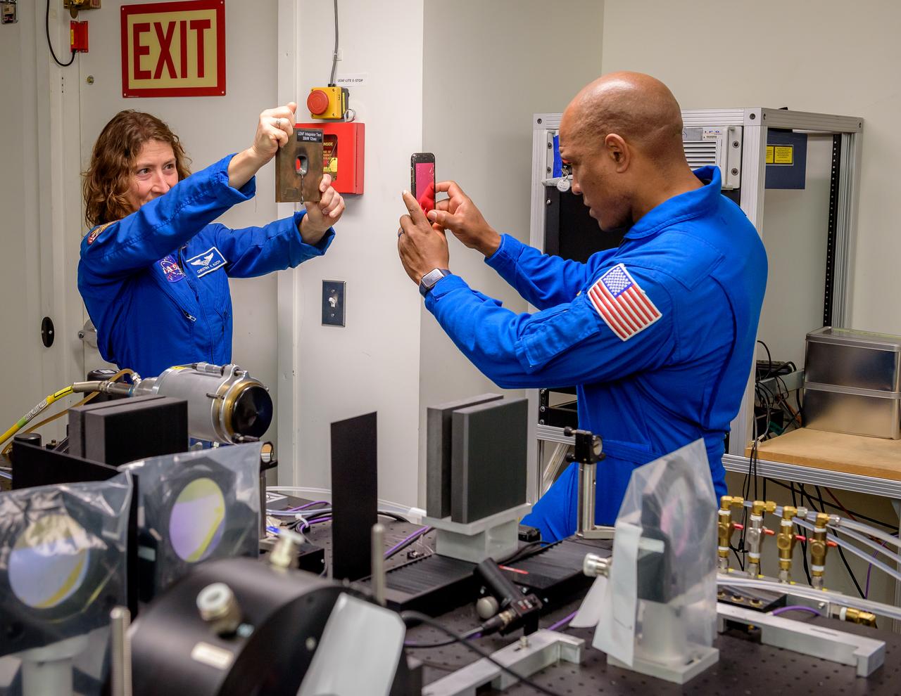 NASA Astronaut Christina Koch, left, holds a test sample for Victor J. Glover to photograph.  The sample is a half-inch steel plate with a hole that was drilled by a 12-second burst from a 30kW laser in the Laser Enhanced Arc Jet Facility (LEAF) laboratory, N238.